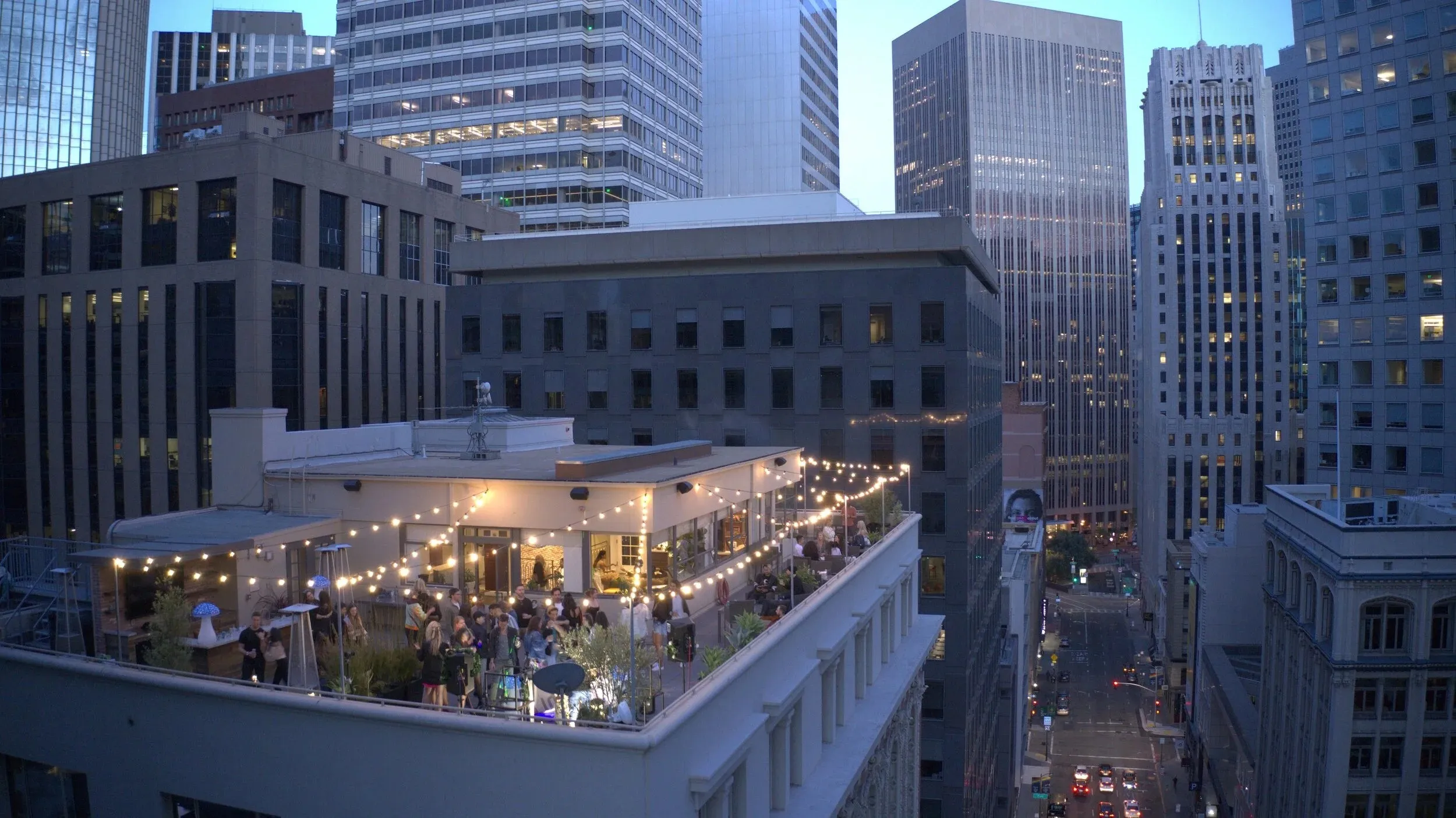 Rooftop party at dusk with string lights and a crowd, surrounded by tall city buildings, creating a lively urban atmosphere.
