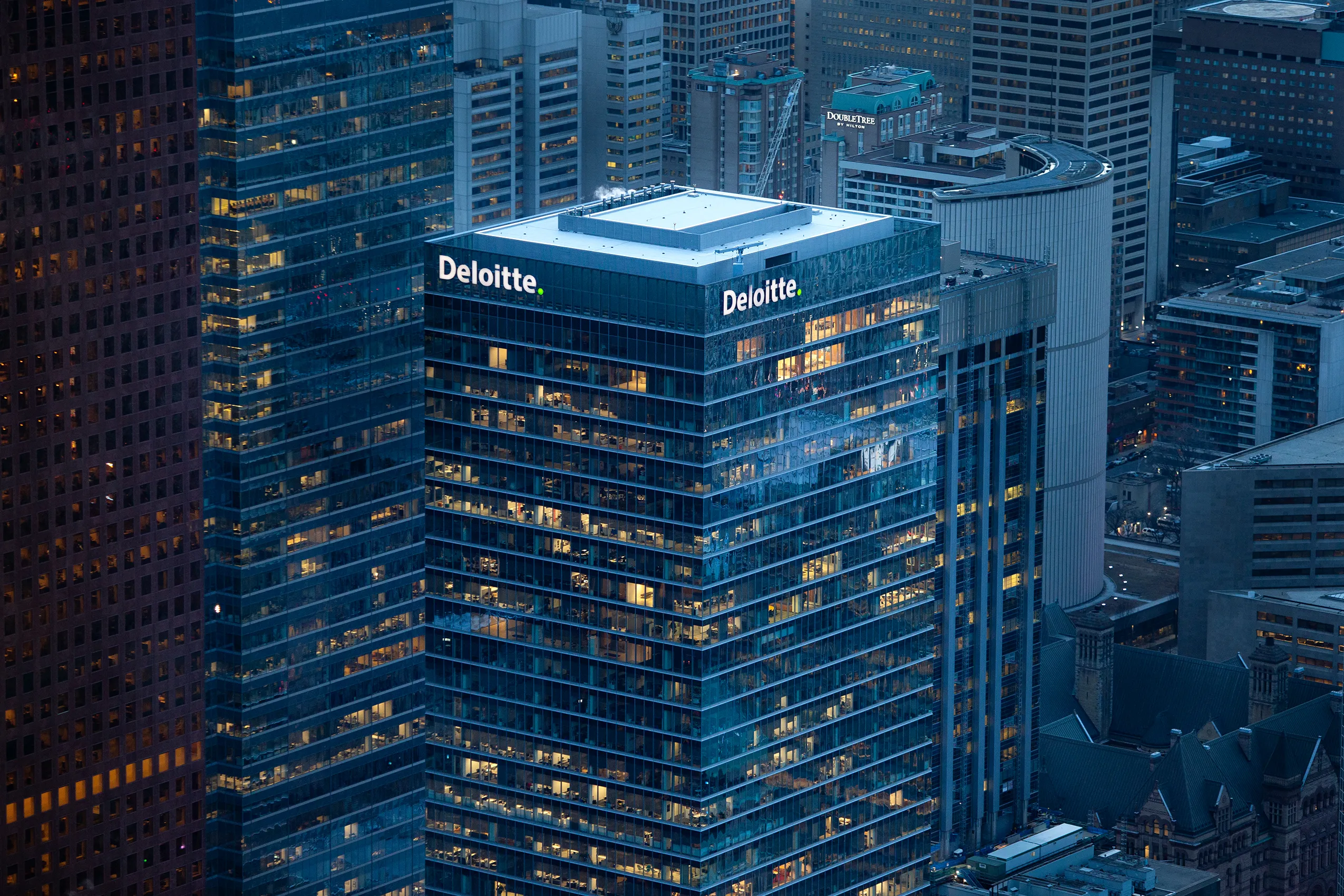 Aerial view of a cityscape at dusk with a modern glass skyscraper lit up, displaying the Deloitte logo at the top on two sides.