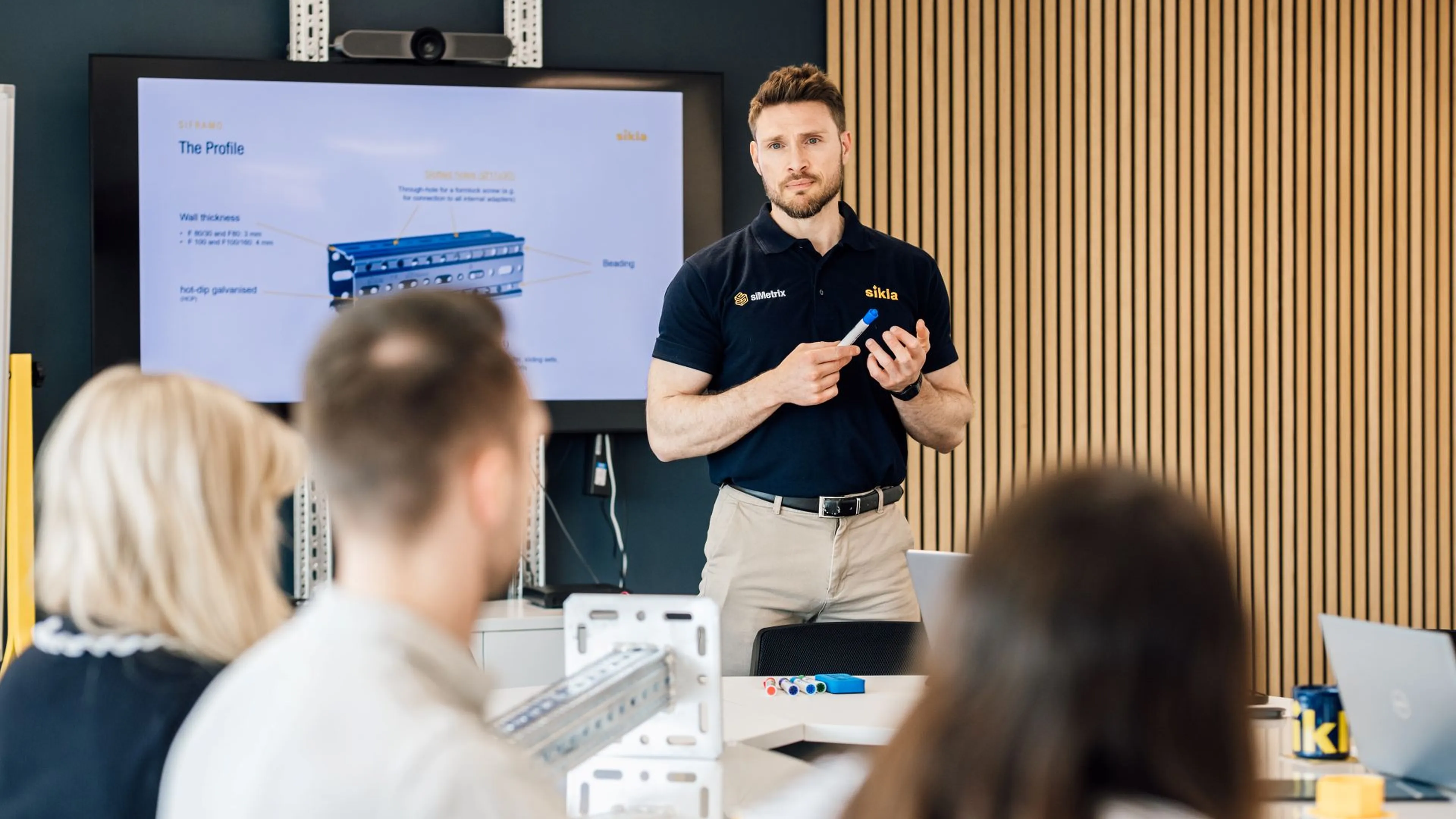 Man in navy polo holding a marker presenting a technical diagram to seated colleagues in a modern meeting room.