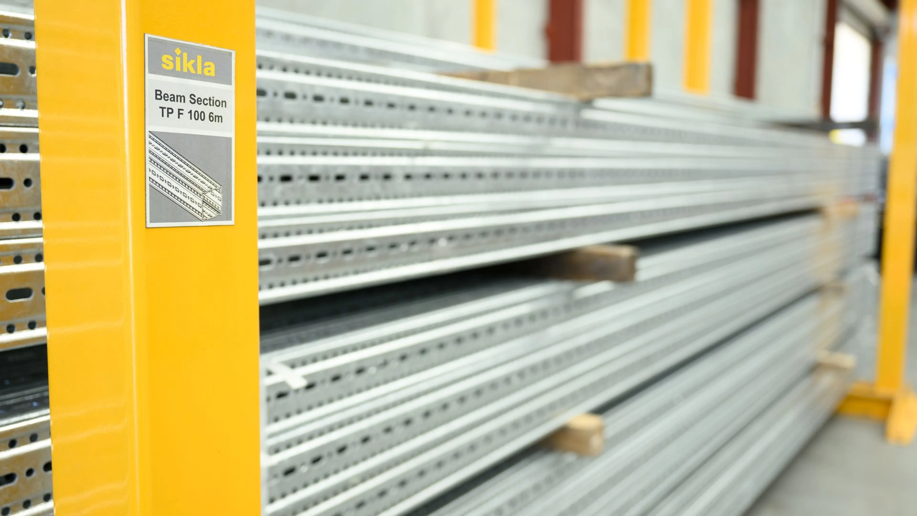 Stacked steel beams on yellow shelving in a warehouse, with a label reading "sikla Beam Section TP F 100 6m" on the left.