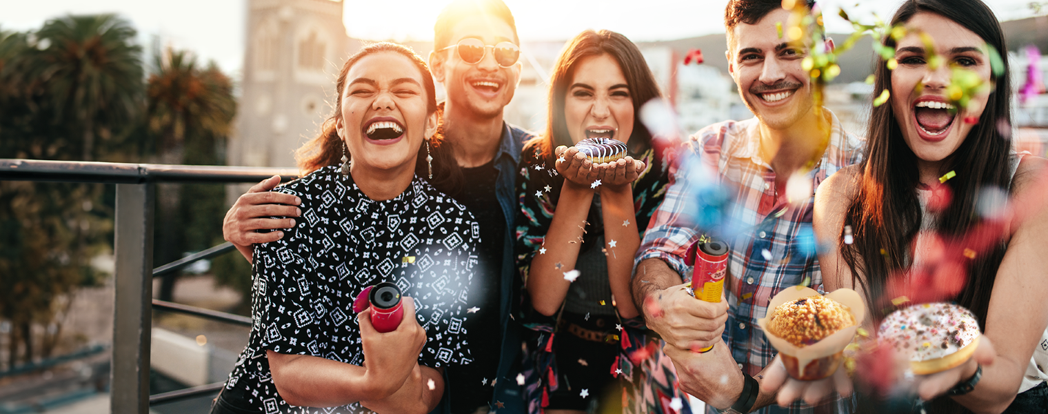 Group of friends celebrating outdoors, smiling and holding donuts and confetti poppers, with a cityscape in the background.