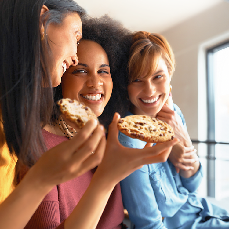 Three women smiling and holding cookies, enjoying a casual moment together indoors, with natural light streaming through a window.