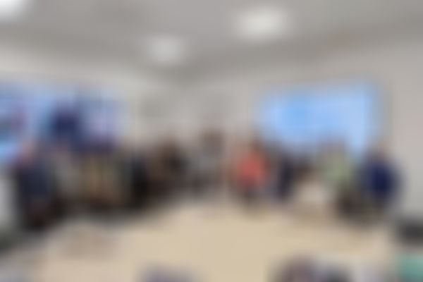 A group of people posing in a conference room, with laptops and a presentation screen in the background. Video call participants are visible on a screen.