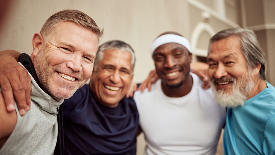 Vier lachende Männer in Sportkleidung umarmen sich für ein Selfie im Freien.