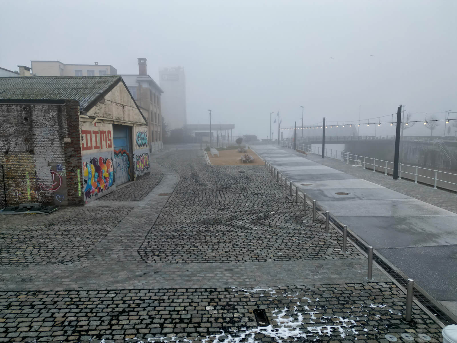 Foggy urban street with cobblestones, graffiti on a building, and string lights. Distant buildings and a misty atmosphere.