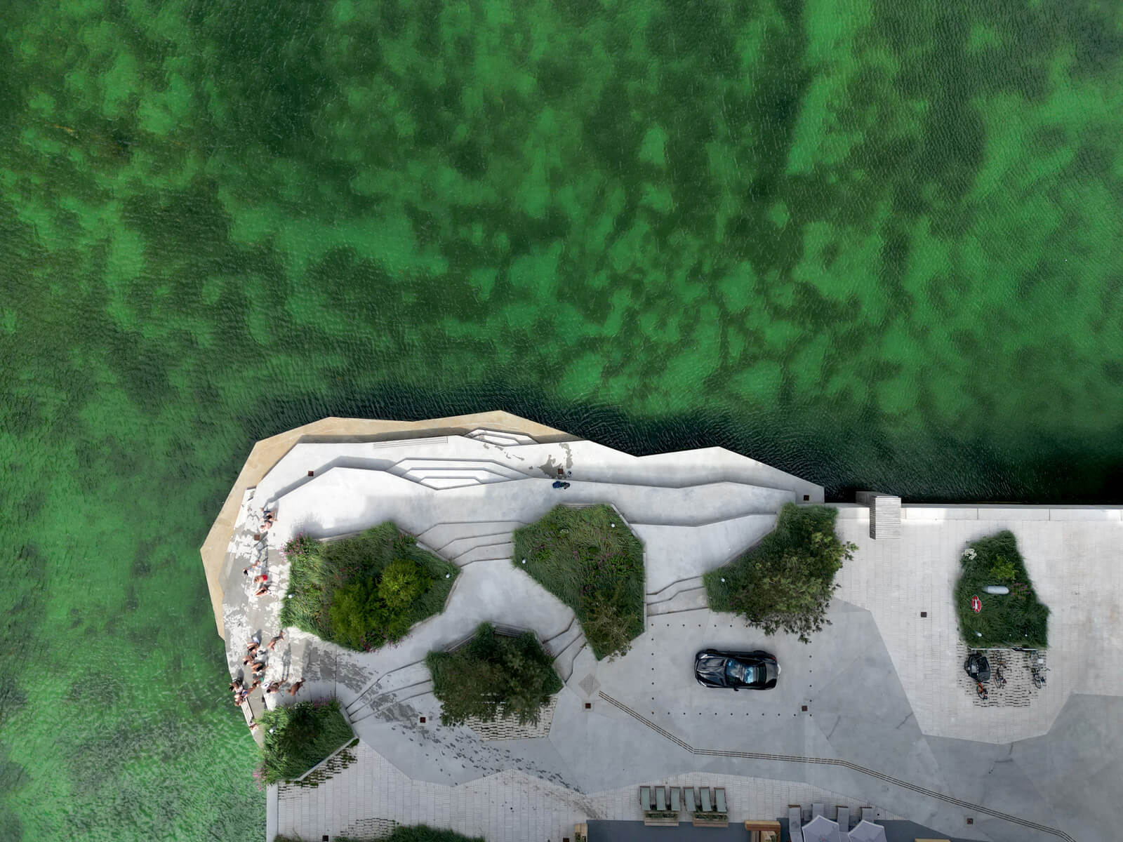 Aerial view of a waterfront area with a zigzagging pathway, trees, a parked car, and green water on the left.