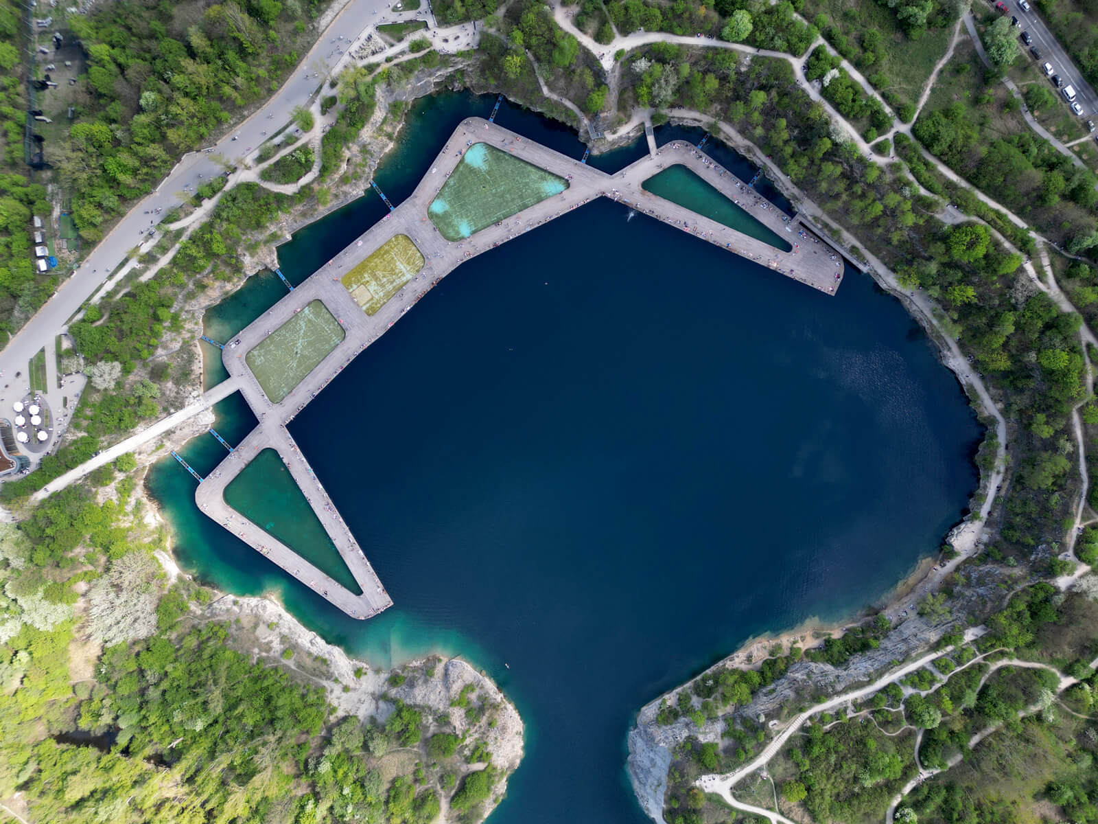Aerial view of a large, irregularly shaped blue lake surrounded by green trees and winding paths, with geometric platforms extending into the water.