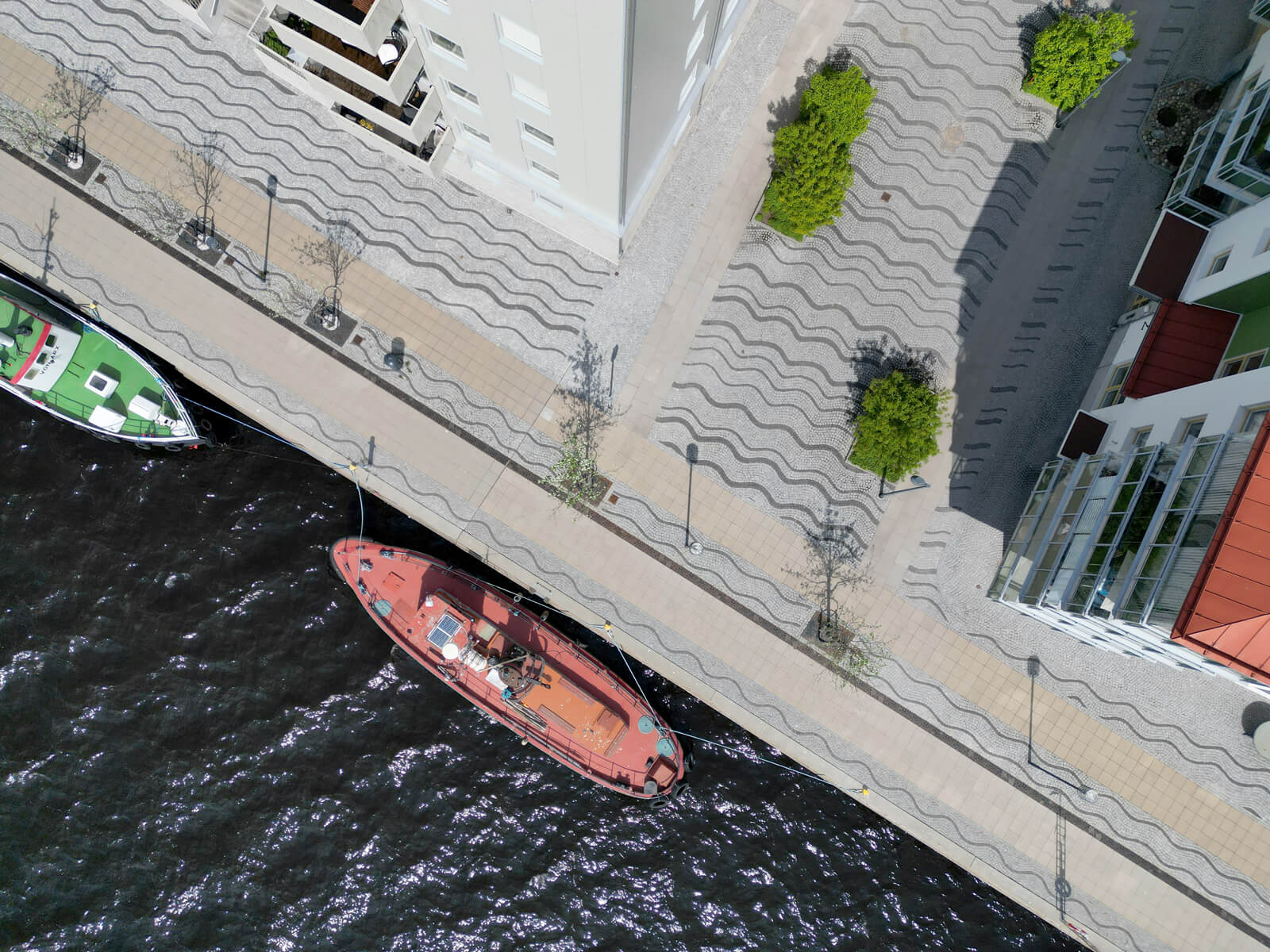 Aerial view of two boats docked alongside a wavy-patterned promenade with trees and modern buildings.