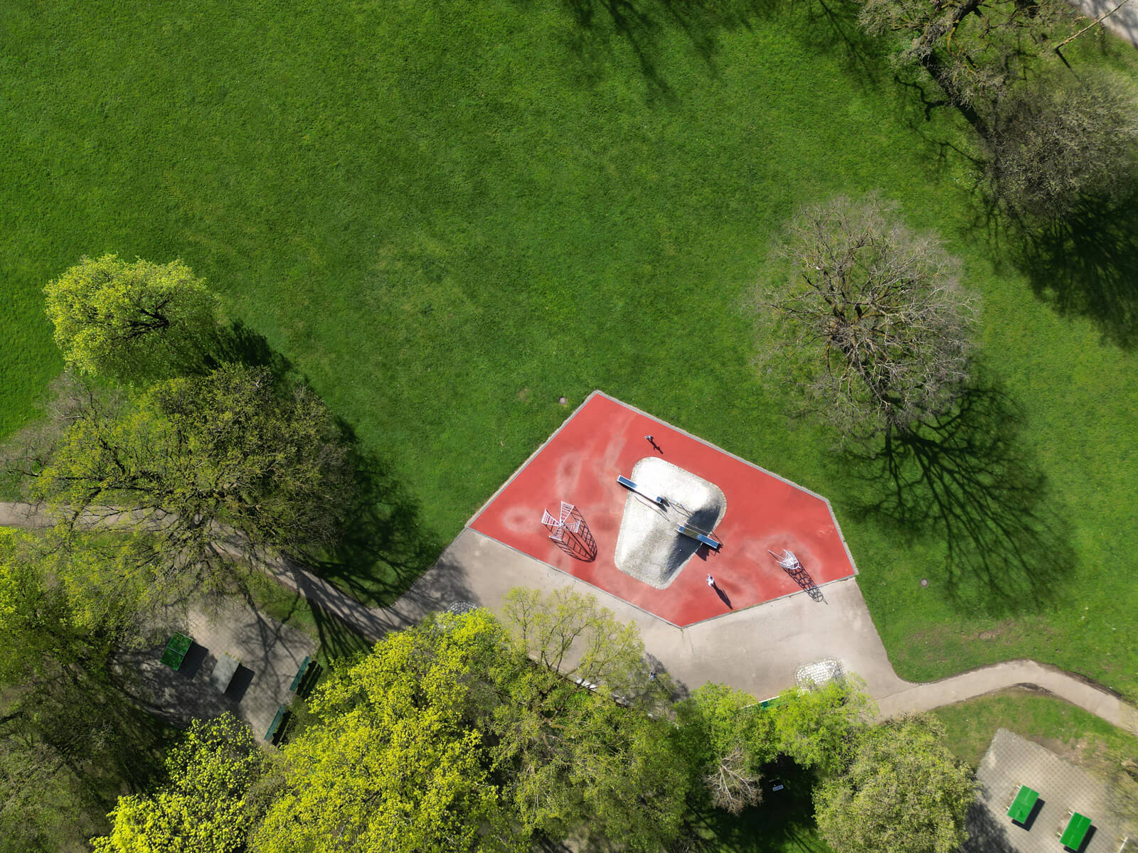 Aerial view of a small playground with a red surface, slide, and climbing structure, surrounded by green grass and trees.