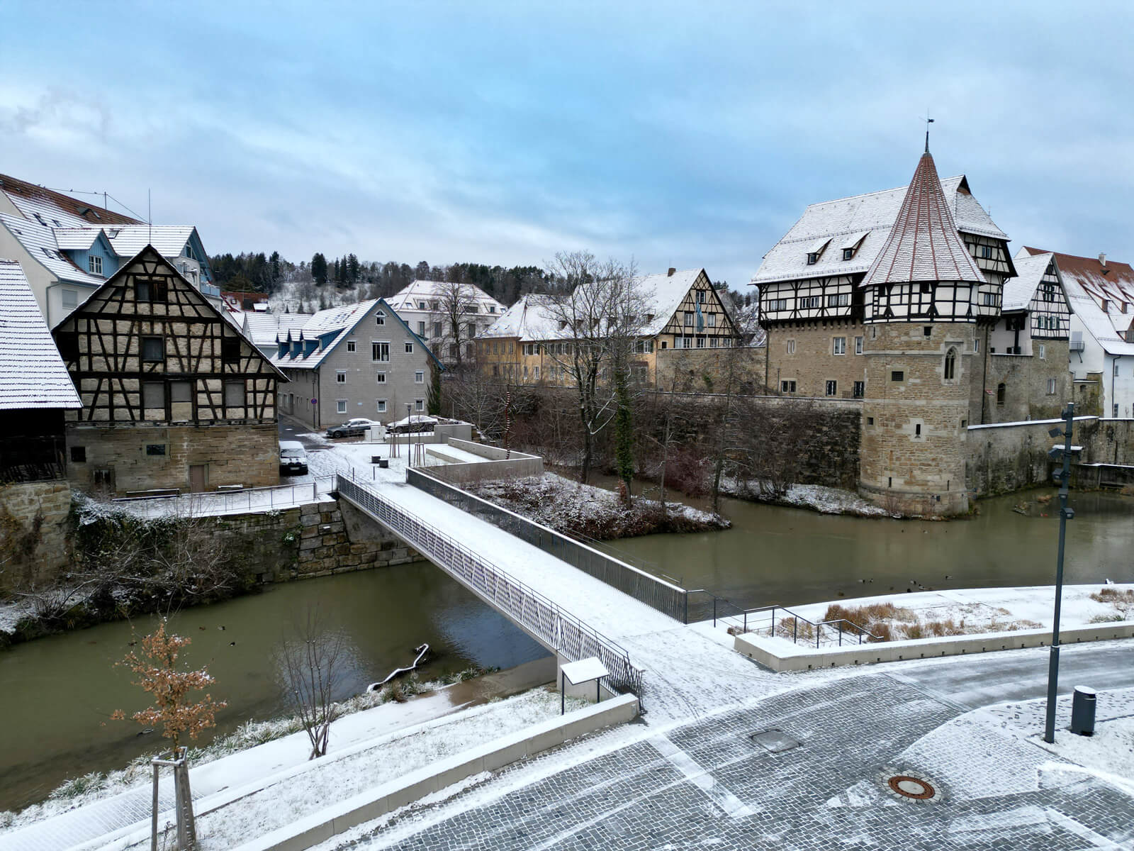 Snow-covered medieval buildings and a bridge over a river, surrounded by a quaint village landscape under a cloudy sky.