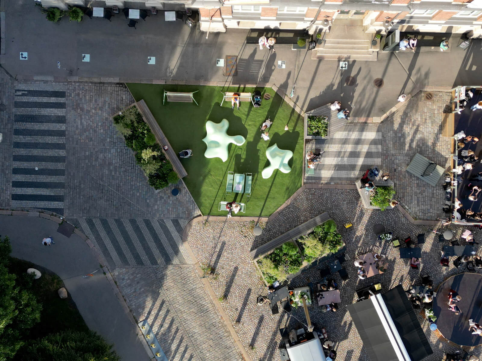 Aerial view of a geometric urban park with green spaces, benches, and people gathered around tables on a cobblestone plaza.