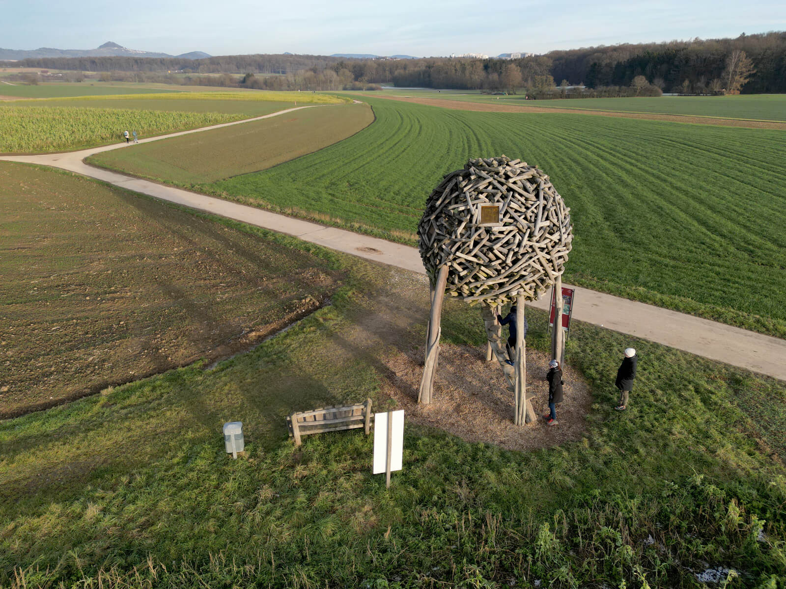 Aerial view of a spherical wooden structure on stilts by a rural path, surrounded by green fields and a few people nearby.