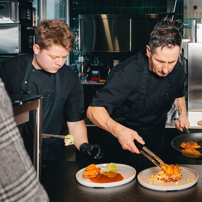 Two chefs in black uniforms plating gourmet dishes in a professional kitchen, focusing intently on presentation.