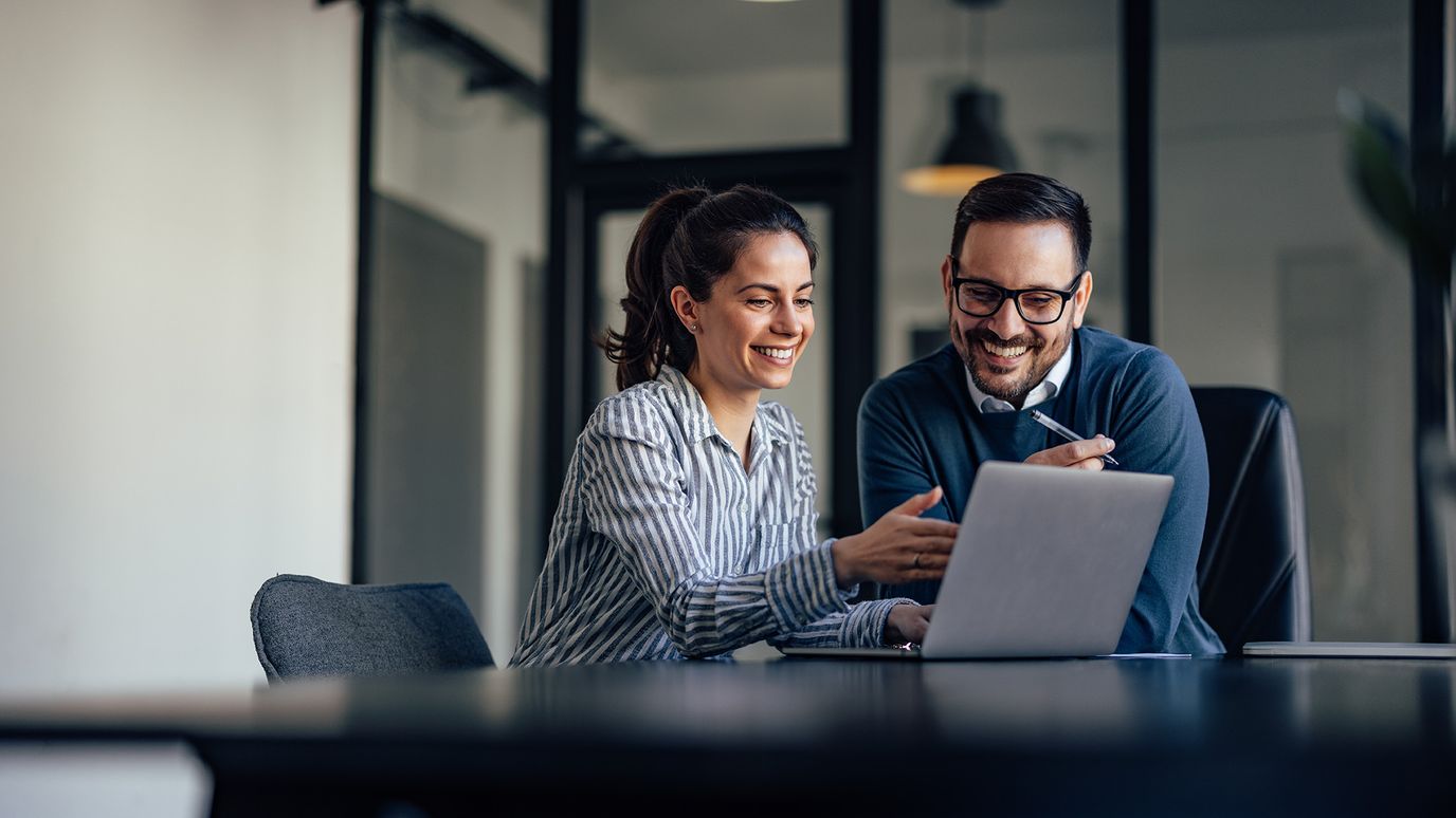 Two colleagues smiling and discussing work while looking at a laptop in a modern office setting.