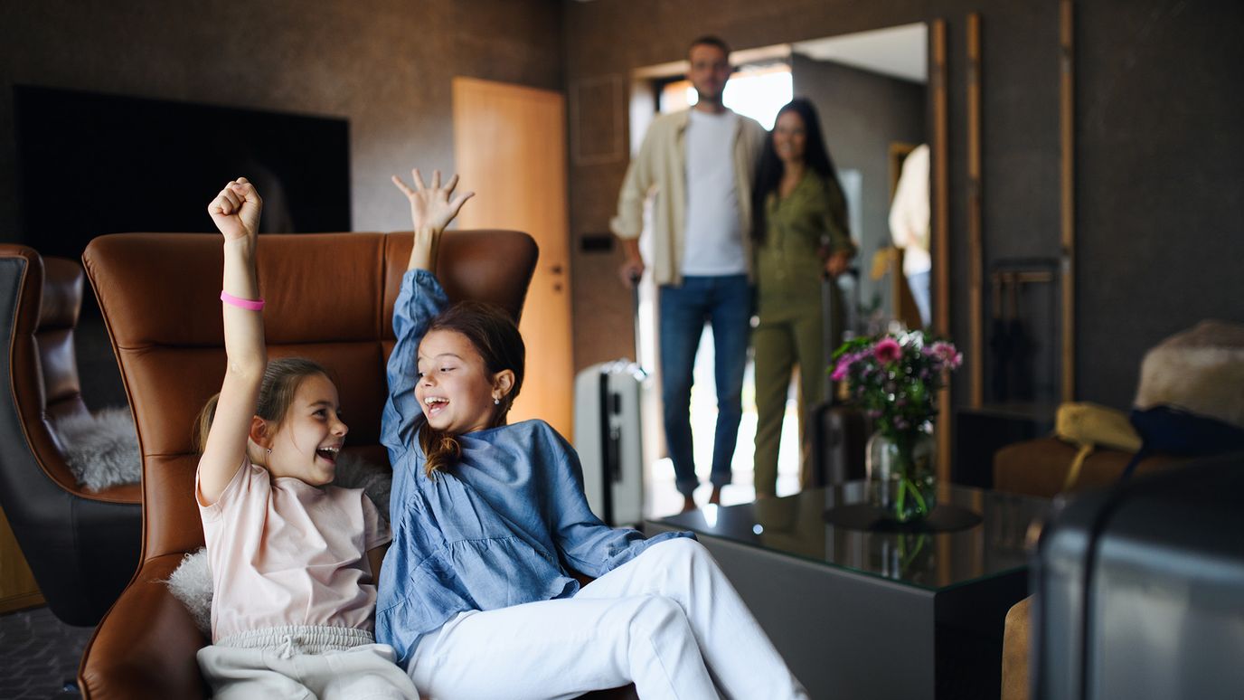 Two excited girls with raised arms sit on chairs in a living room. A smiling couple stands in the background near a door.