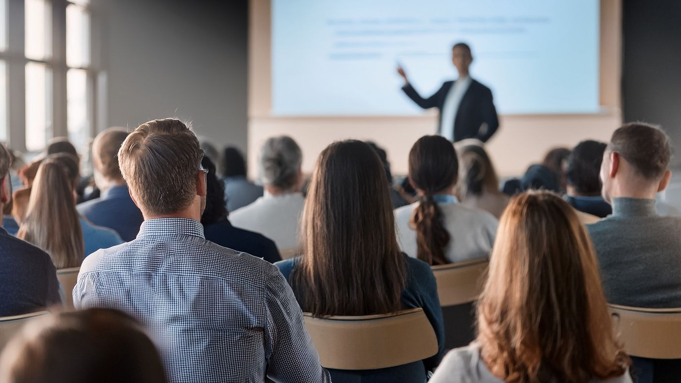 Audience listening to a speaker in a conference room. The speaker stands near a screen displaying a presentation.