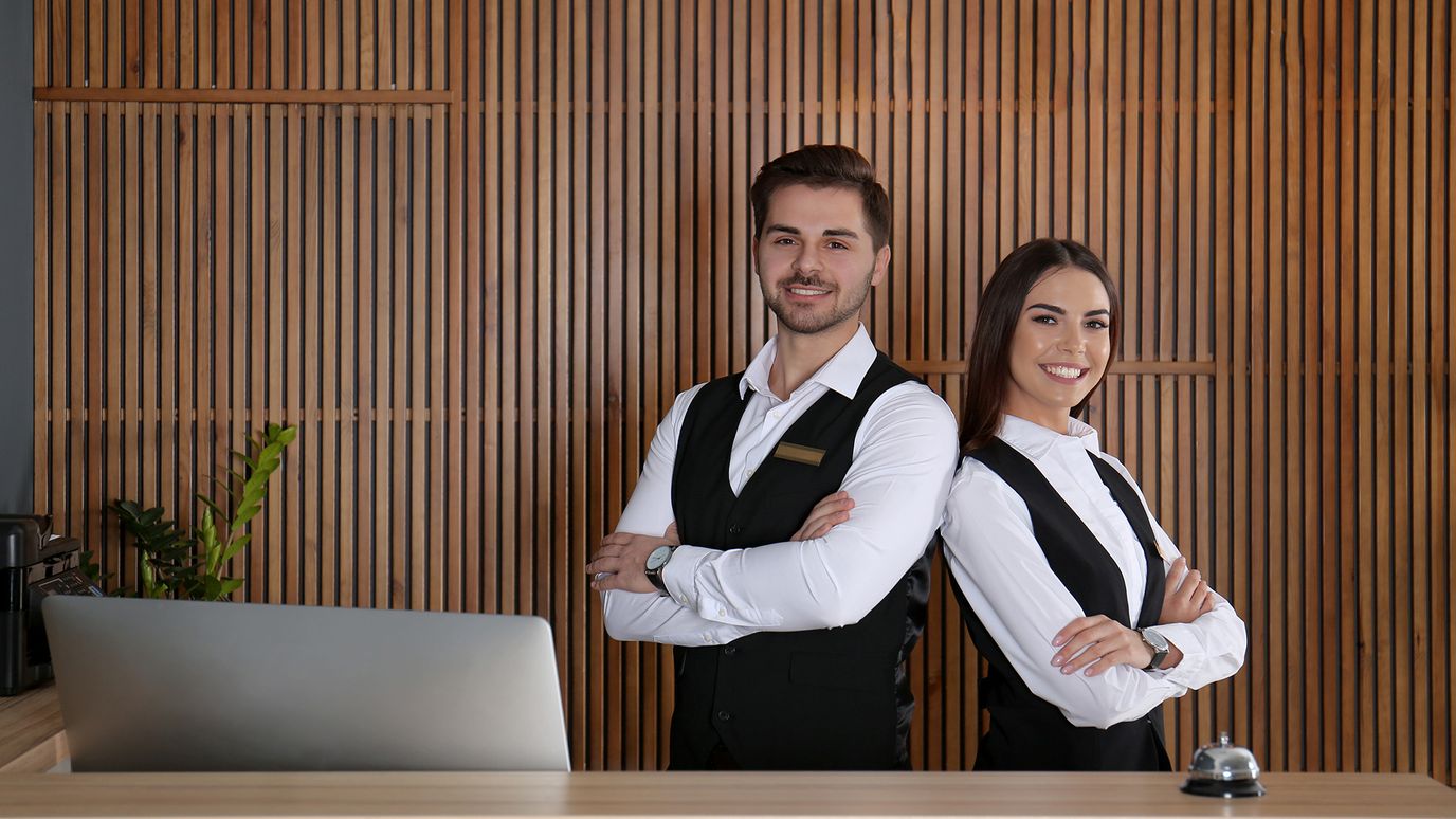 Two smiling receptionists in vests stand with arms crossed at a wooden desk, with a laptop and bell in front.