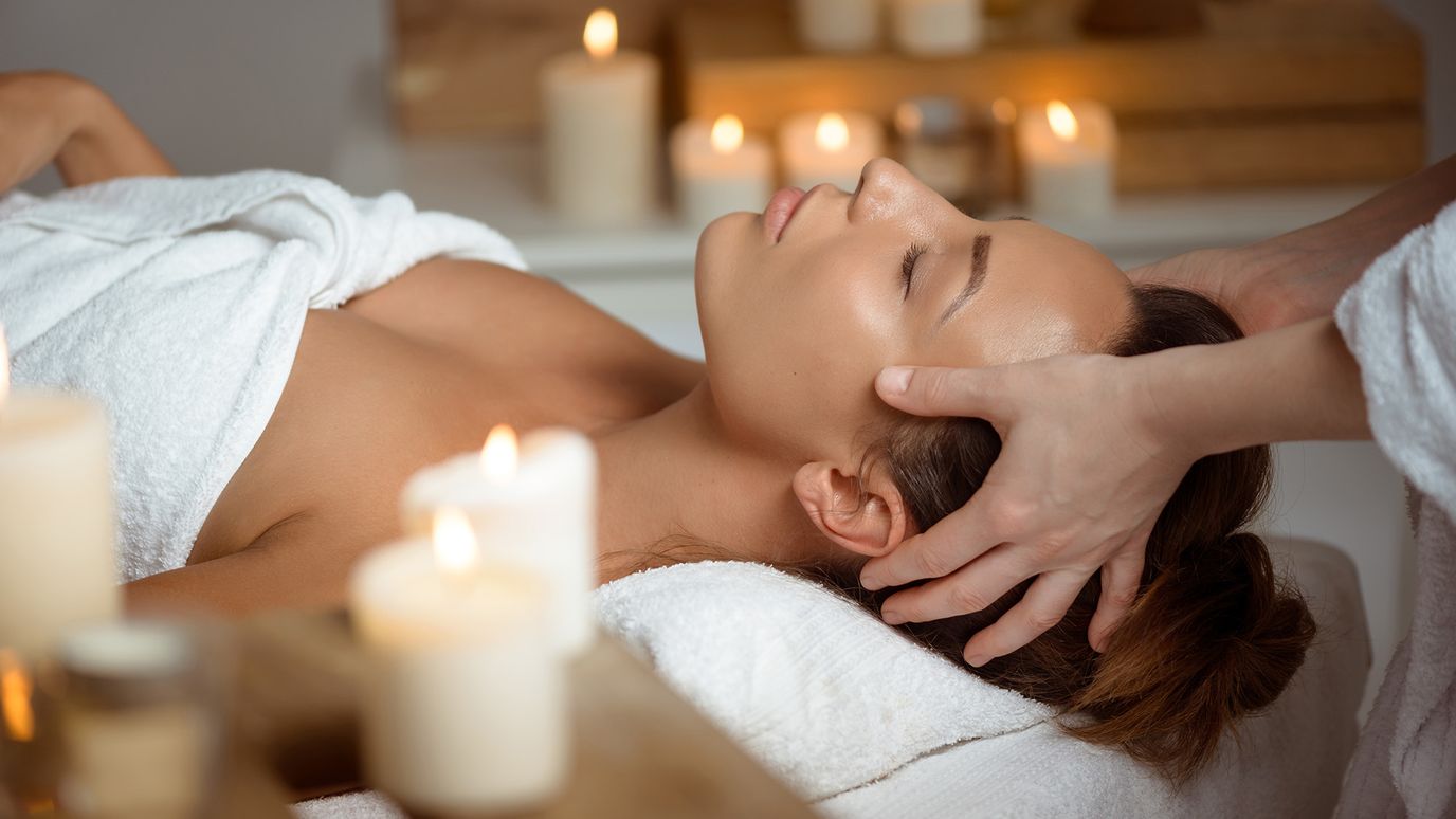 Person receiving a relaxing head massage in a spa setting, surrounded by lit candles and wrapped in a white towel.