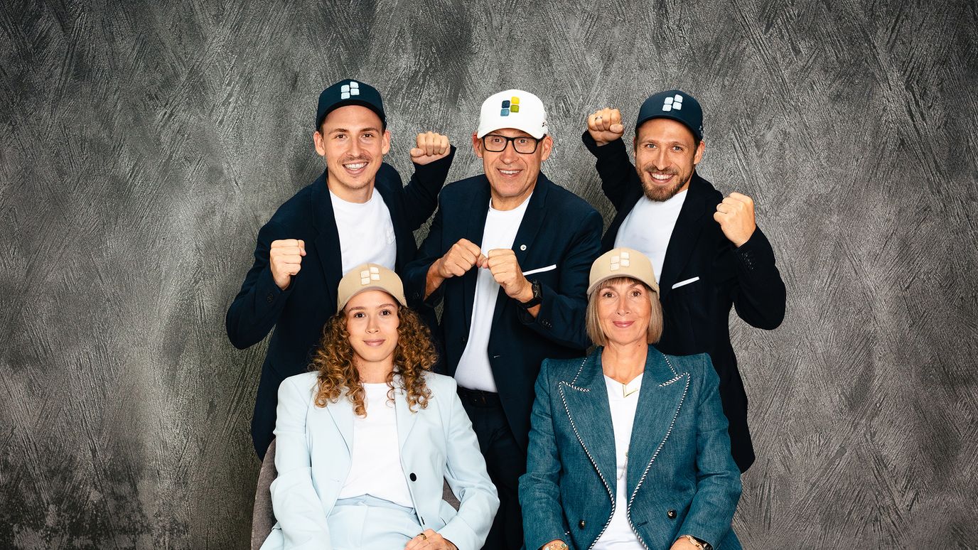 Five people in coordinated suits and caps pose against a textured background, with three standing and two seated, all smiling energetically.