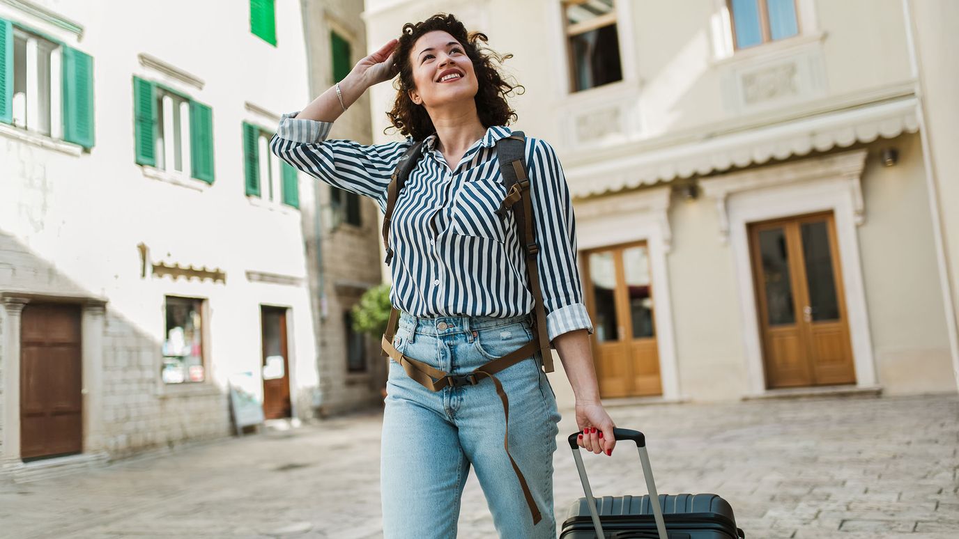 Woman in striped shirt and jeans, smiling while pulling a suitcase, with historic buildings in the background.