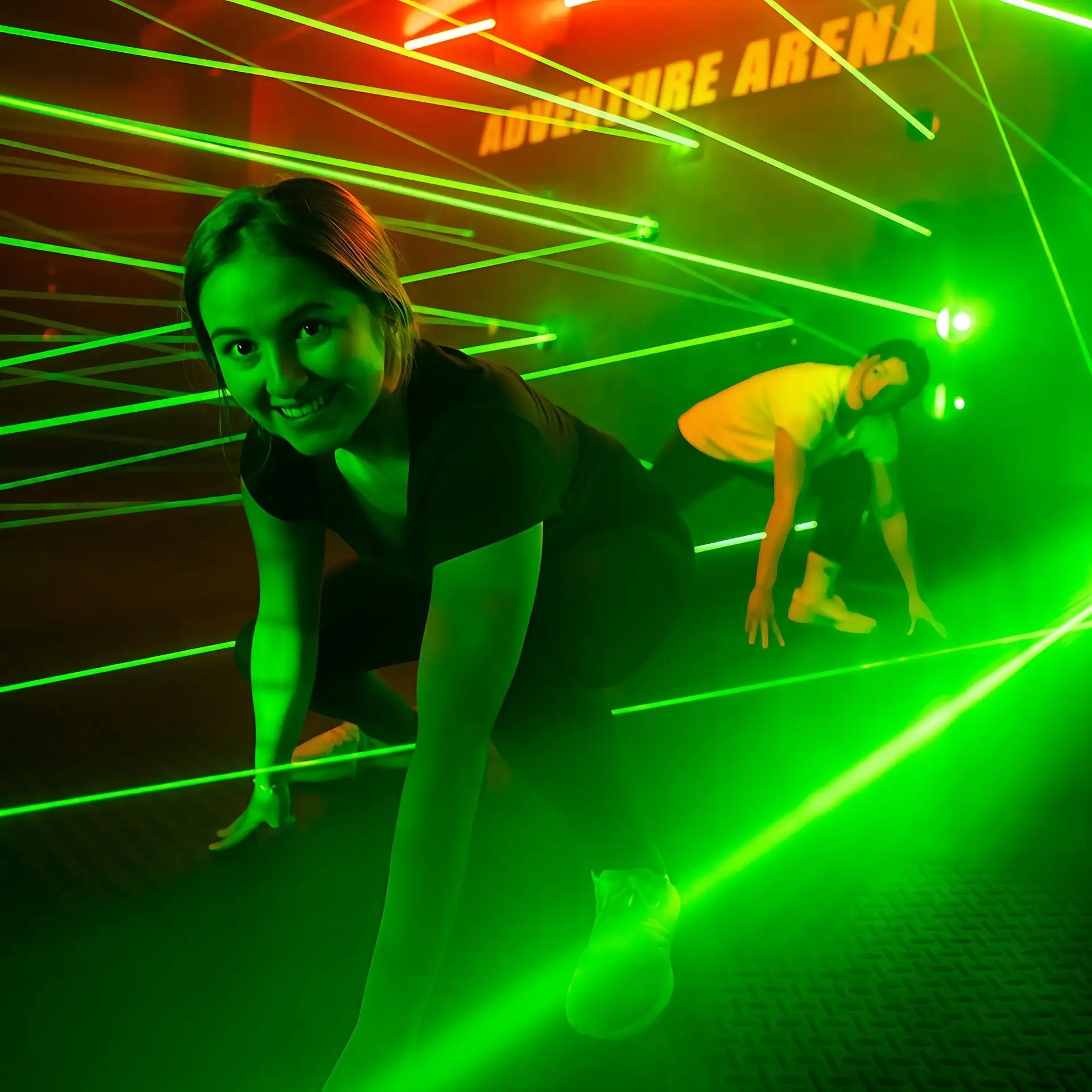 Two people navigate through a maze of green laser beams in a dimly lit room with "Adventure Arena" sign above. Both are crouched and focused.