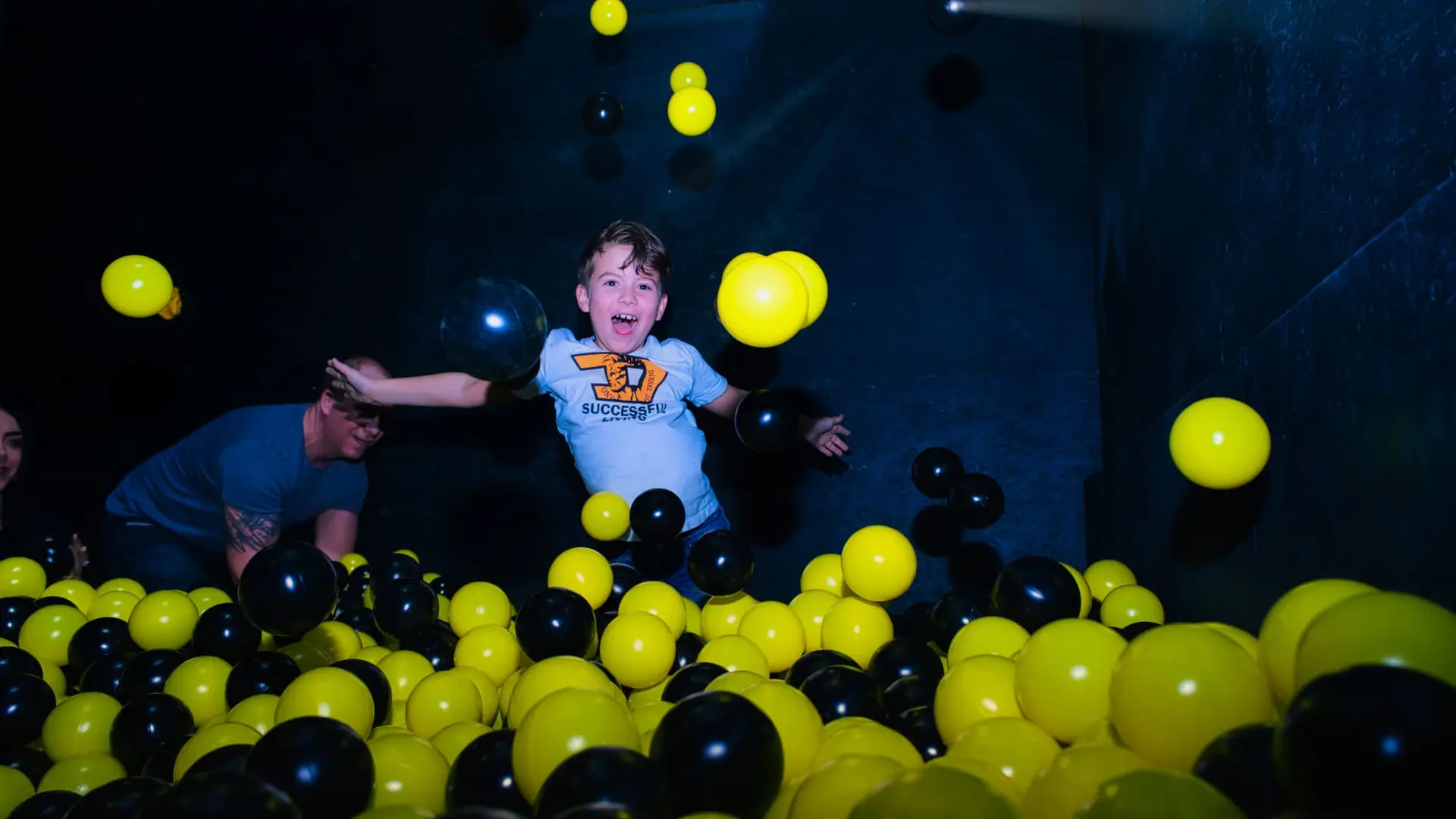 Child joyfully playing in a ball pit filled with black and yellow balls, wearing a white T-shirt, in a dimly lit room.