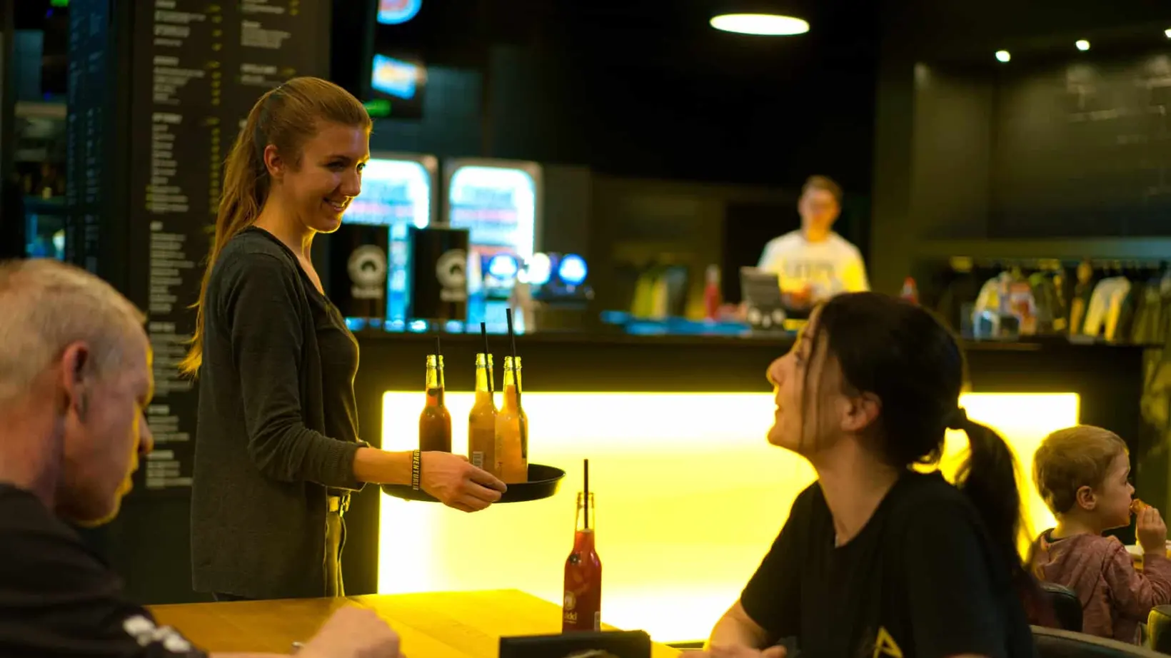 Servicemitarbeiterin serviert Flaschengetränke an einem Tisch im Adventure-Arena-Bistro; Gäste essen und unterhalten sich vor einer gelb beleuchteten Bar.