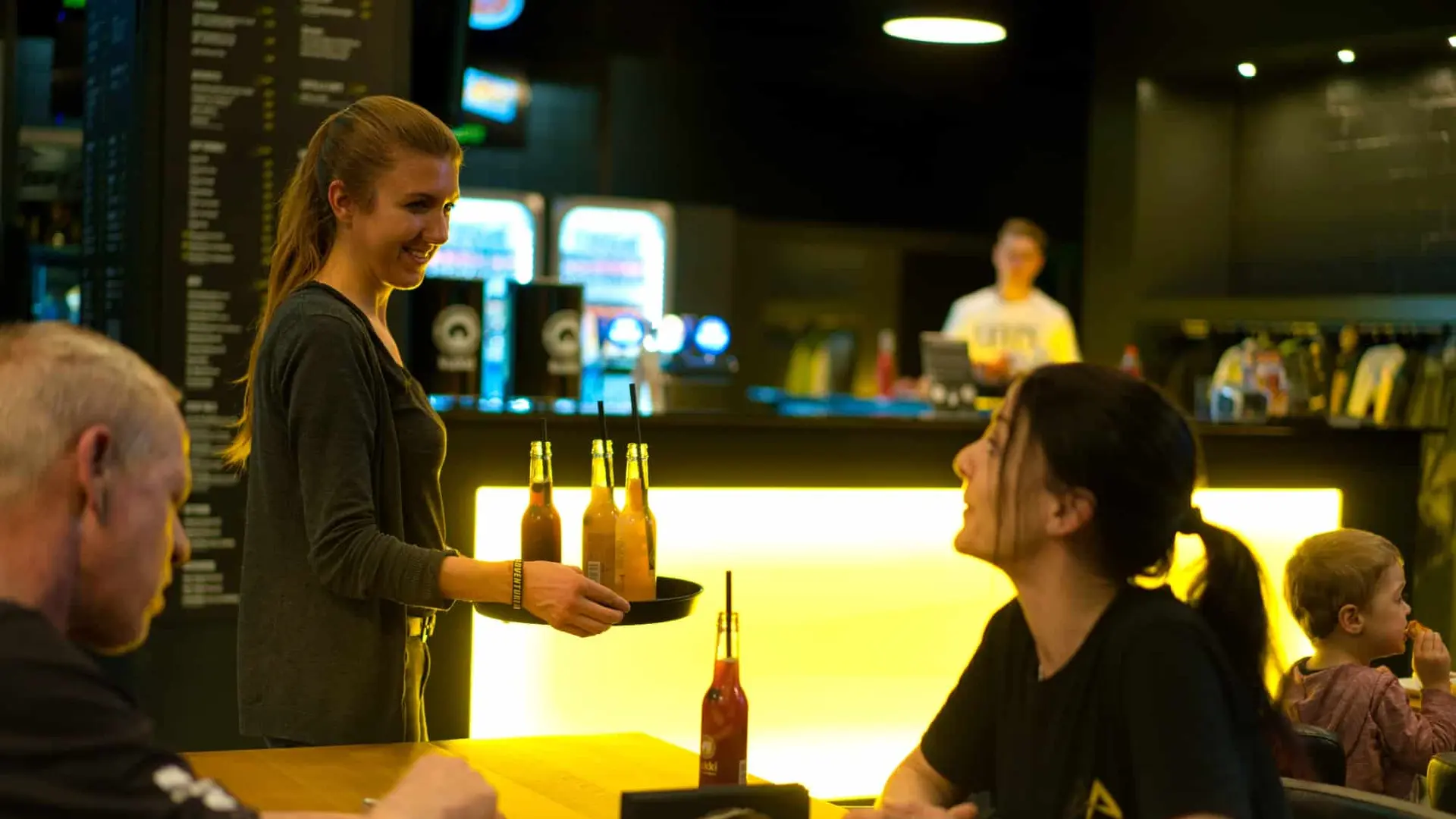 Servicemitarbeiterin serviert Flaschengetränke an einem Tisch im Adventure-Arena-Bistro; Gäste essen und unterhalten sich vor einer gelb beleuchteten Bar.