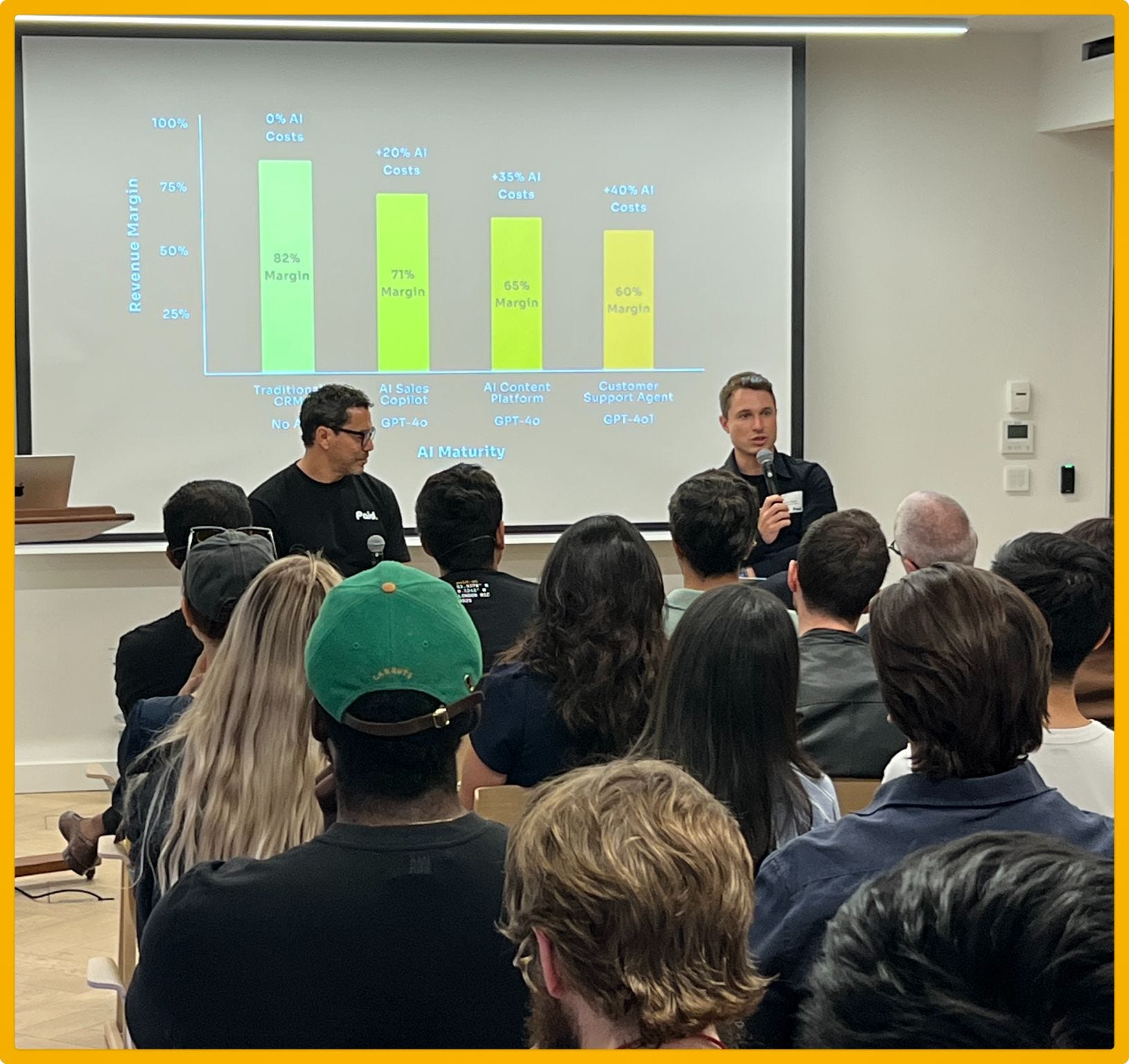 Audience watching two speakers discuss AI costs and margins, with a bar chart displayed on a screen behind them.