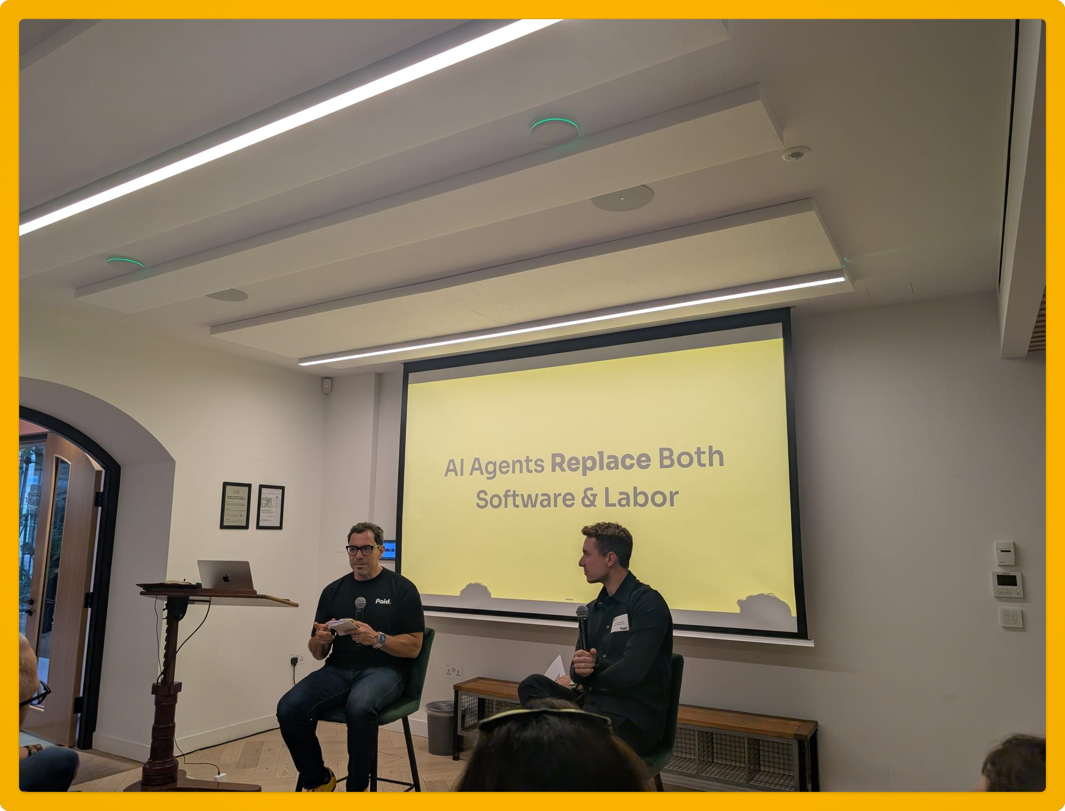 Two people sit on stools in front of a screen displaying 
