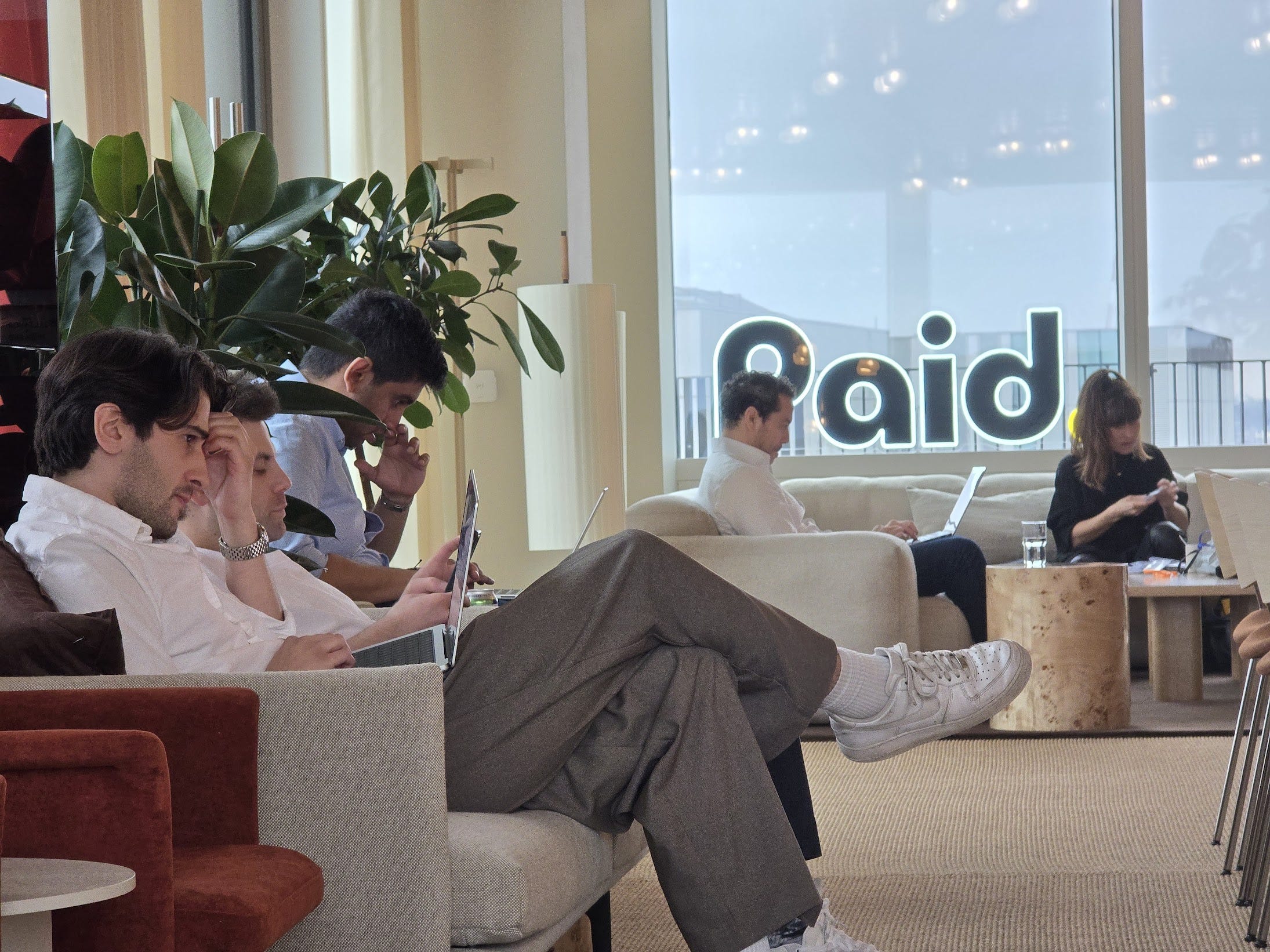 People sitting on couches in a modern lounge, using laptops and phones. Large windows display 