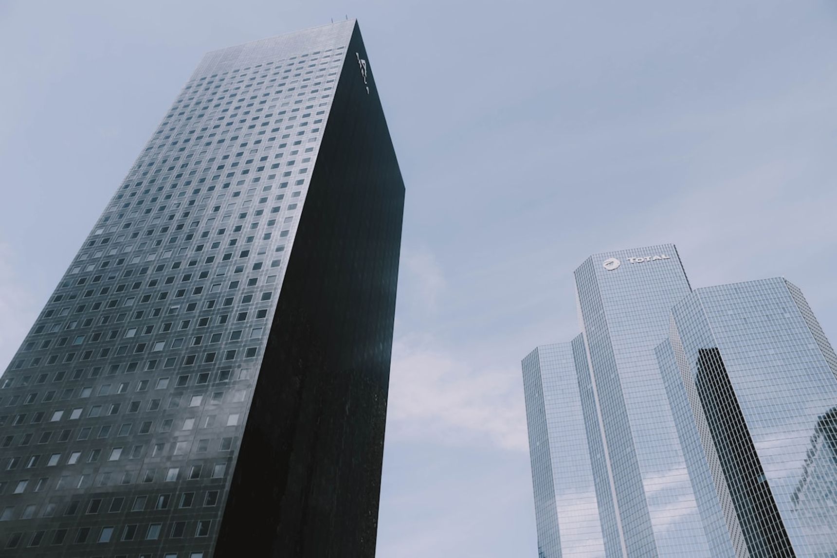 Two modern skyscrapers with reflective glass facades against a clear blue sky. One building is dark, the other is lighter with visible logos.
