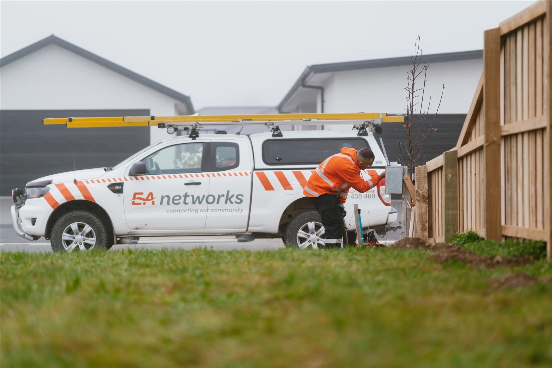 A worker in an orange jacket checks an electrical box near a white utility vehicle marked "EA Networks" with a ladder on top.