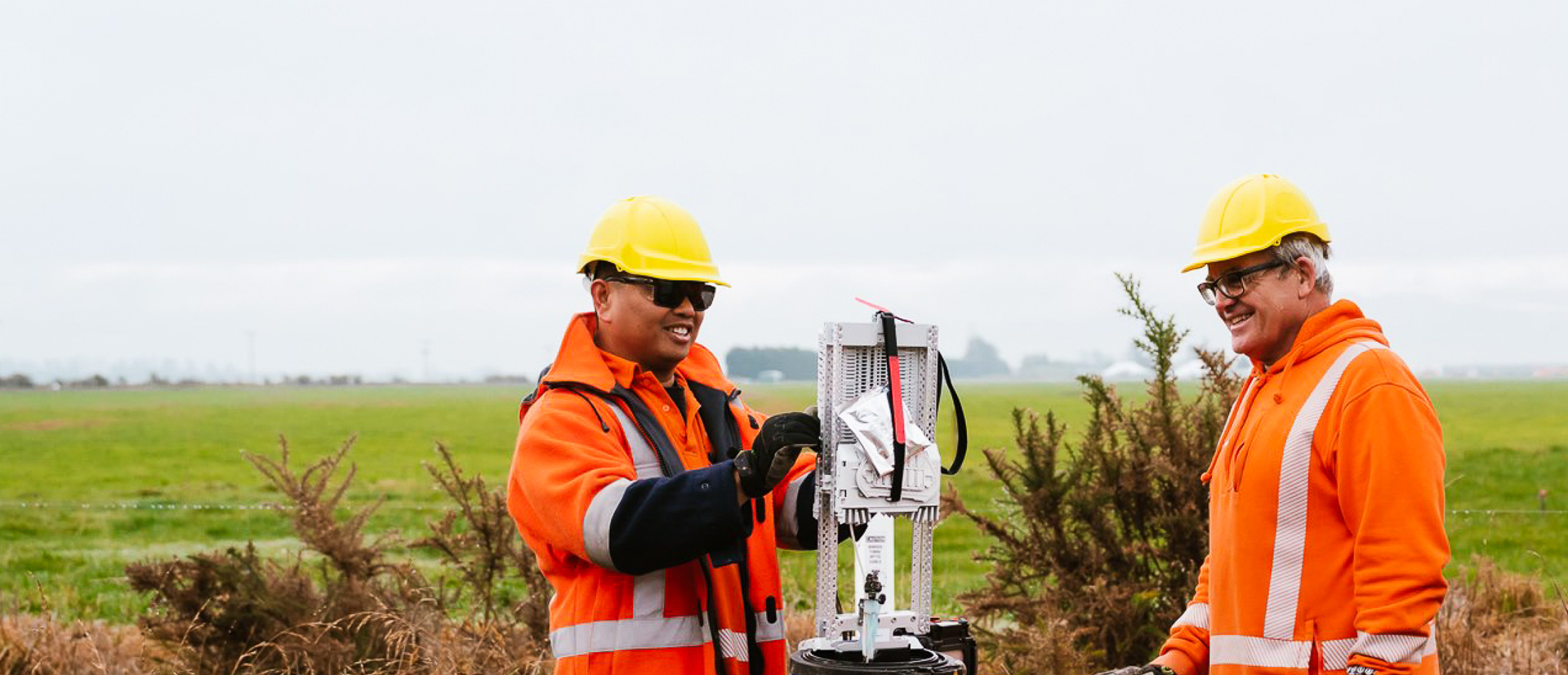 Two workers in orange safety gear and yellow helmets operate equipment in a grassy field, smiling and engaged in their task.