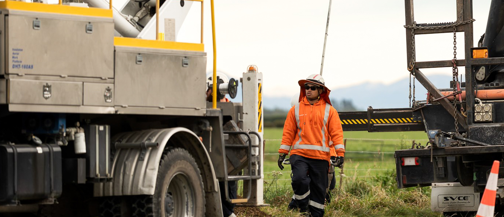 Worker in an orange jacket and hat stands between utility trucks on a grassy field, with mountains in the background.