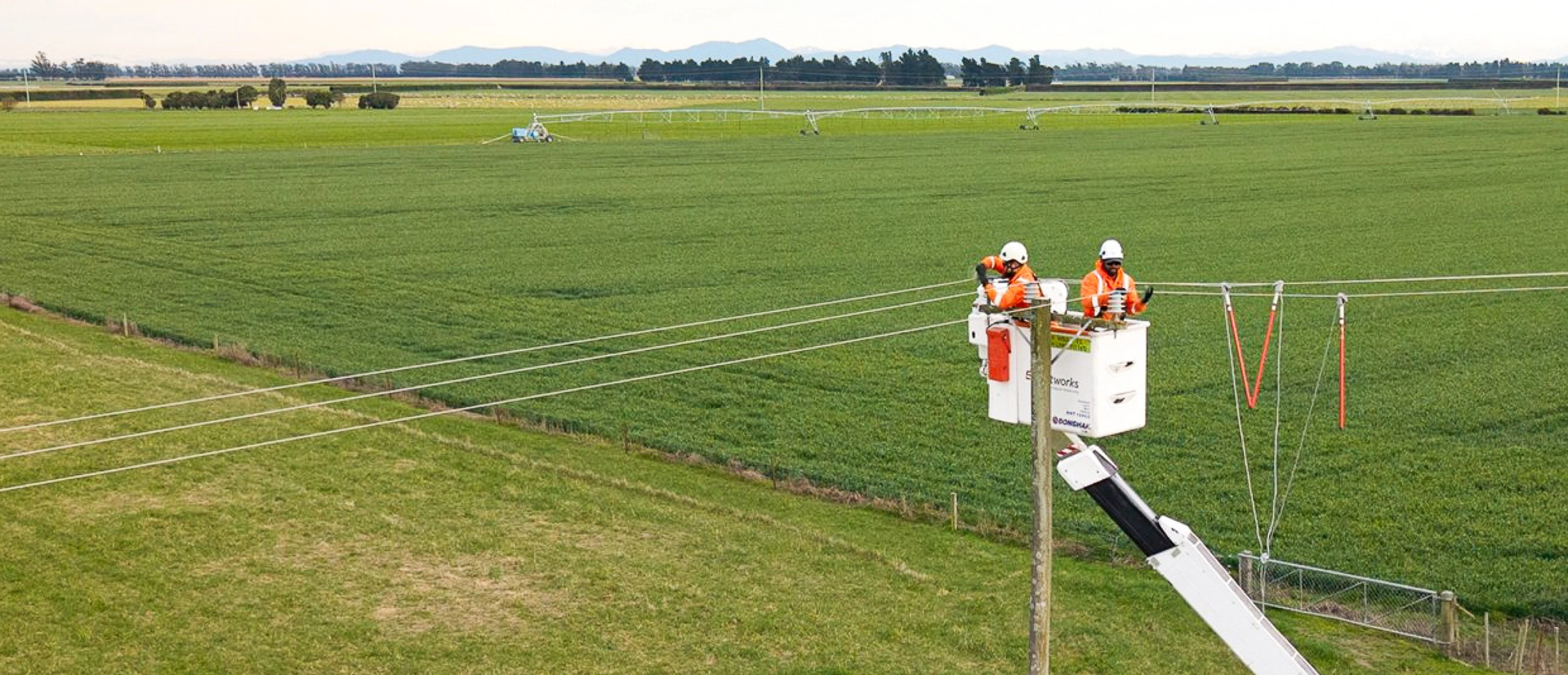 Two workers in a bucket lift repair power lines over a green field, wearing orange safety gear and helmets.