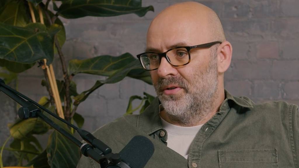 A man wearing glasses speaks into a microphone. A potted plant is behind him.