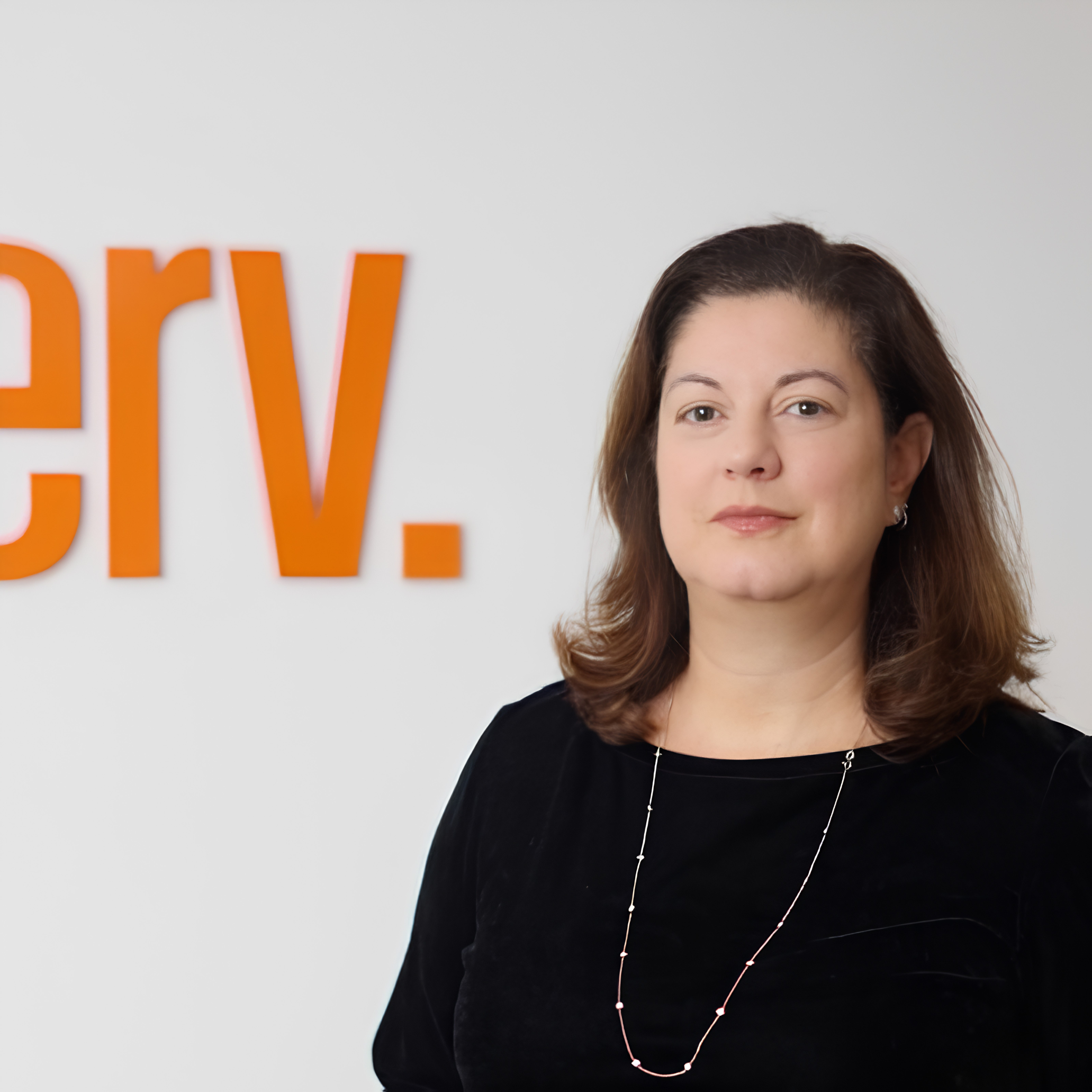 A woman with shoulder-length brown hair wearing a black top and necklace, stands in front of a wall with orange lettering partially visible.