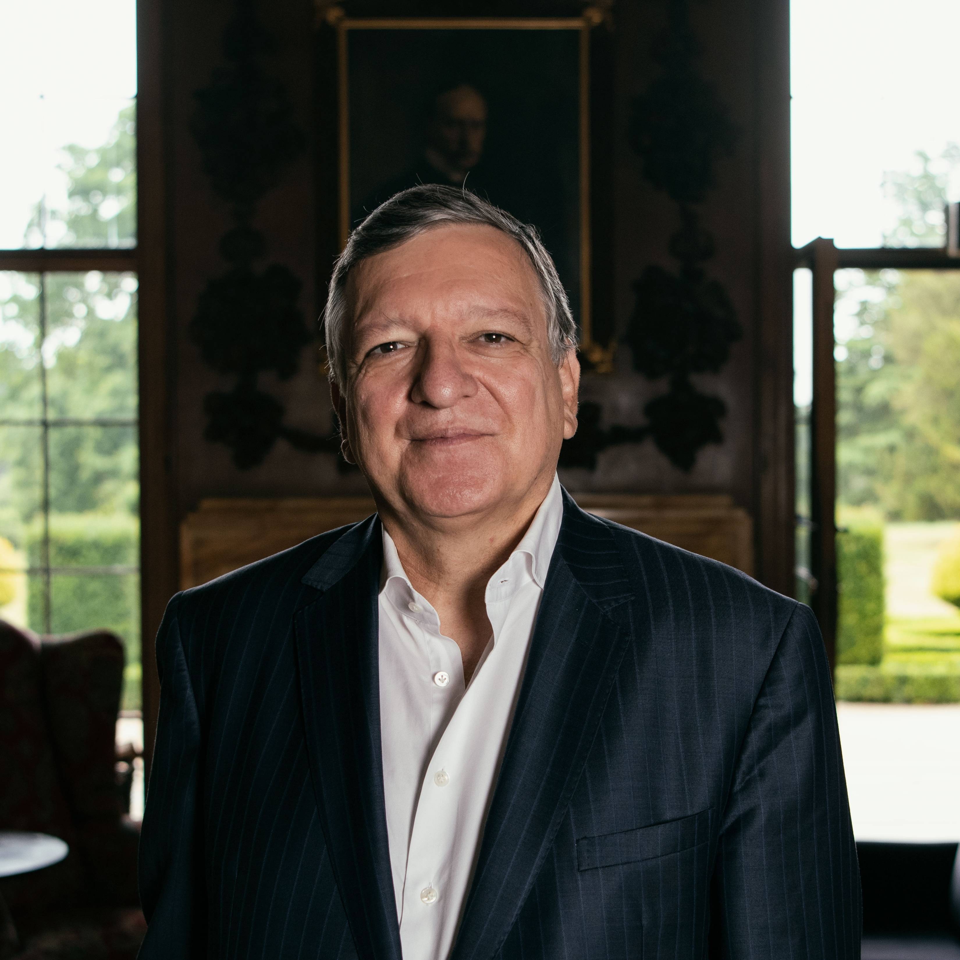 A man in a suit stands indoors, smiling, with a portrait and large windows showing a garden in the background.