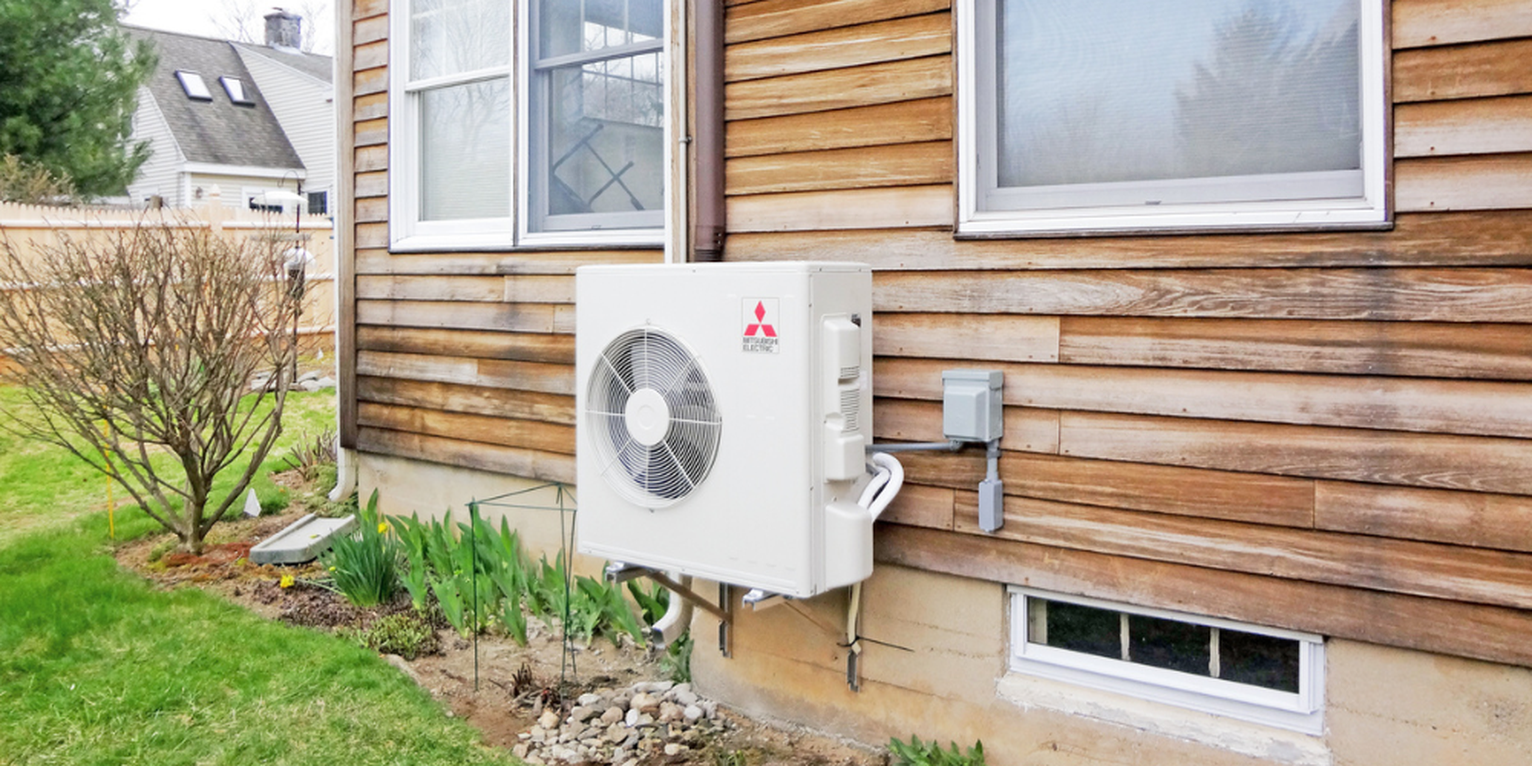 Outdoor air conditioning unit mounted on a wooden house exterior, surrounded by a small garden with green plants.