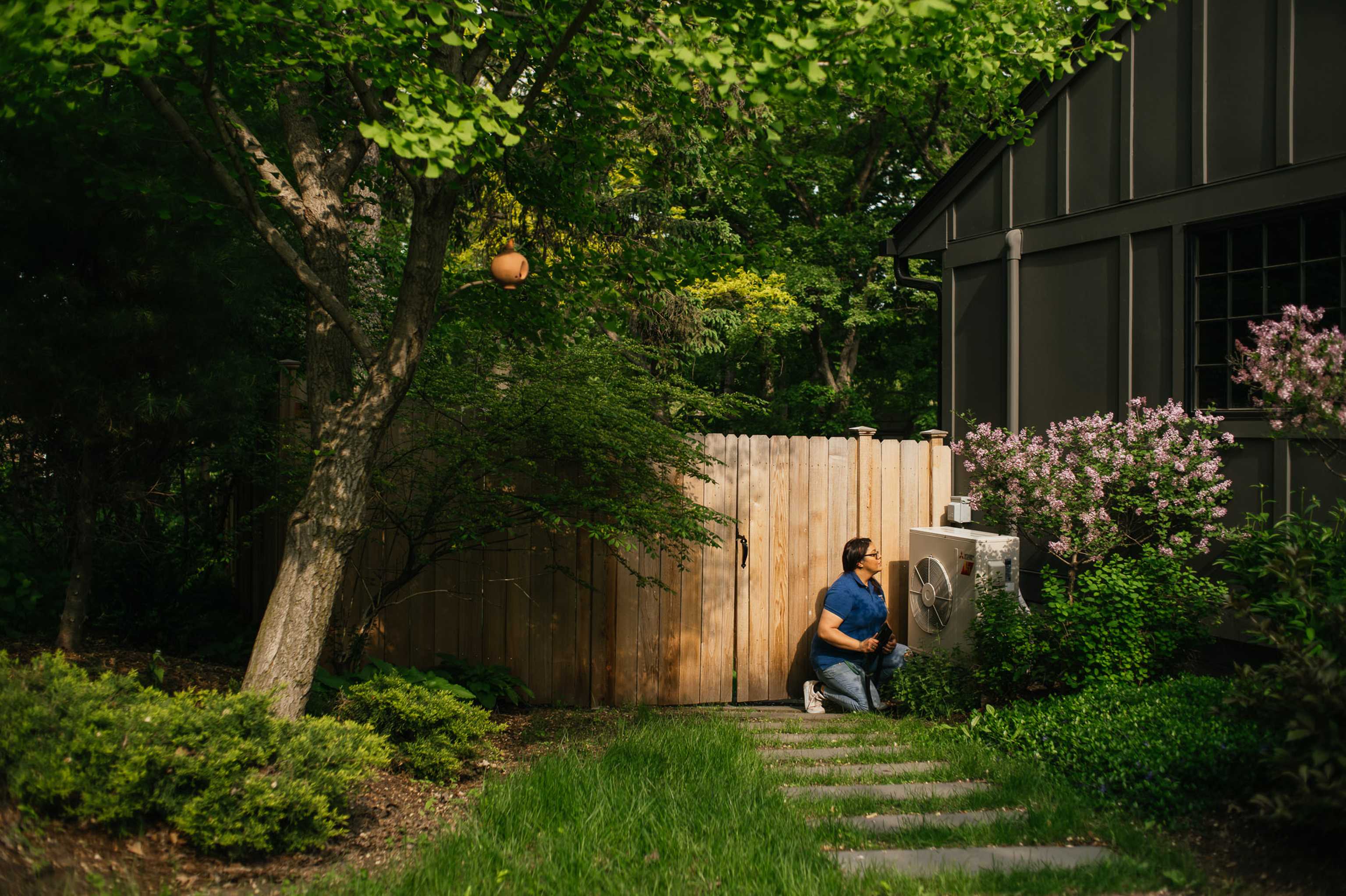Person in blue shirt kneeling by an outdoor air conditioning unit near a wooden fence, surrounded by lush greenery and trees.