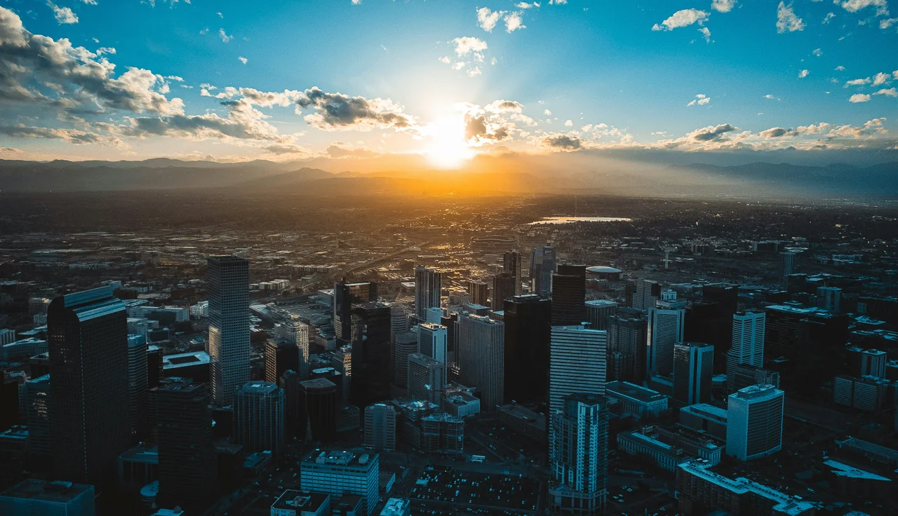 Aerial view of the Denver skyline at sunset, with tall buildings silhouetted against a vibrant sky and sun setting over distant mountains.