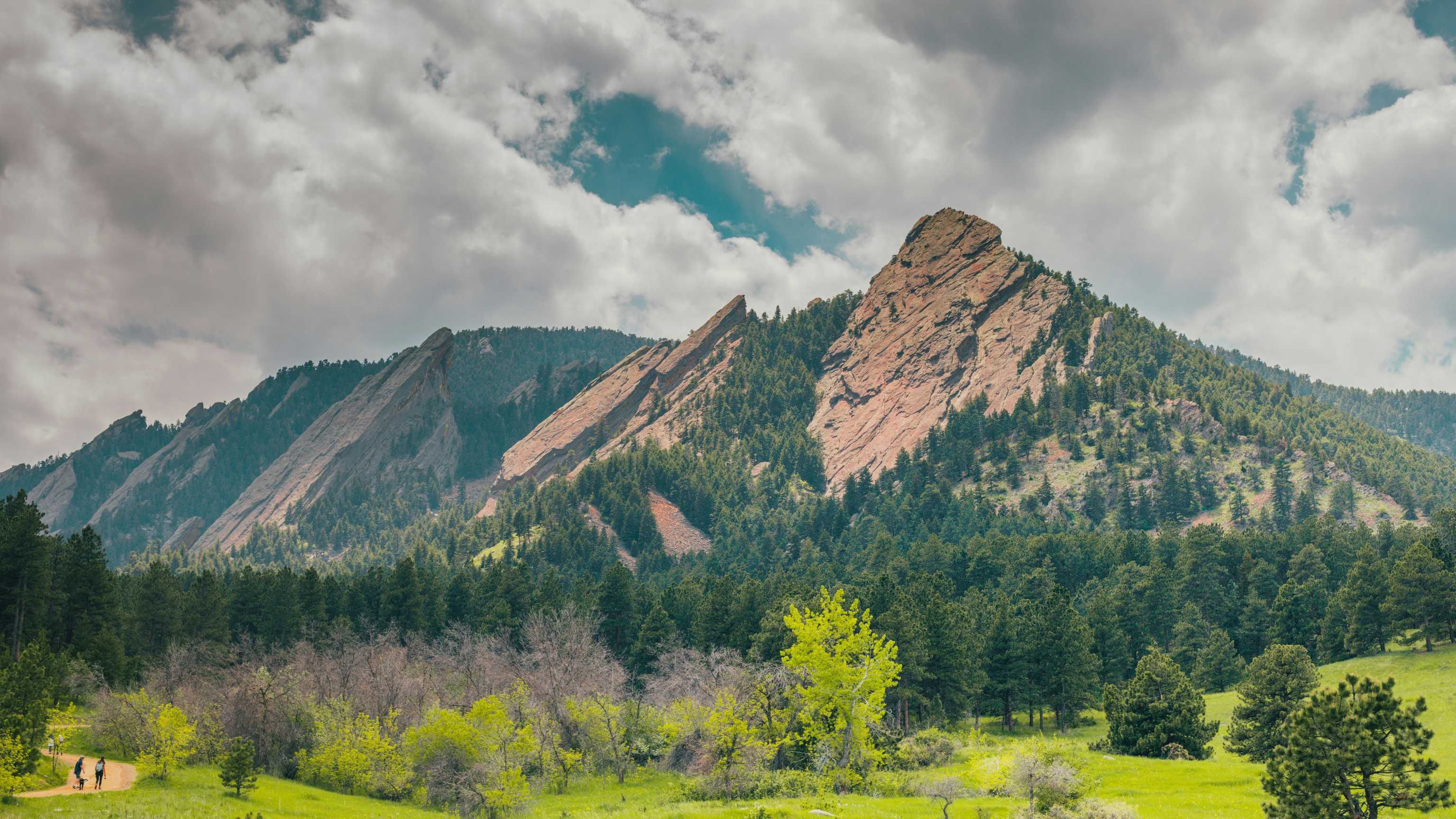 Scenic view of the Flatirons, jagged rock formations in Boulder, Colorado, with lush greenery and a cloudy sky.