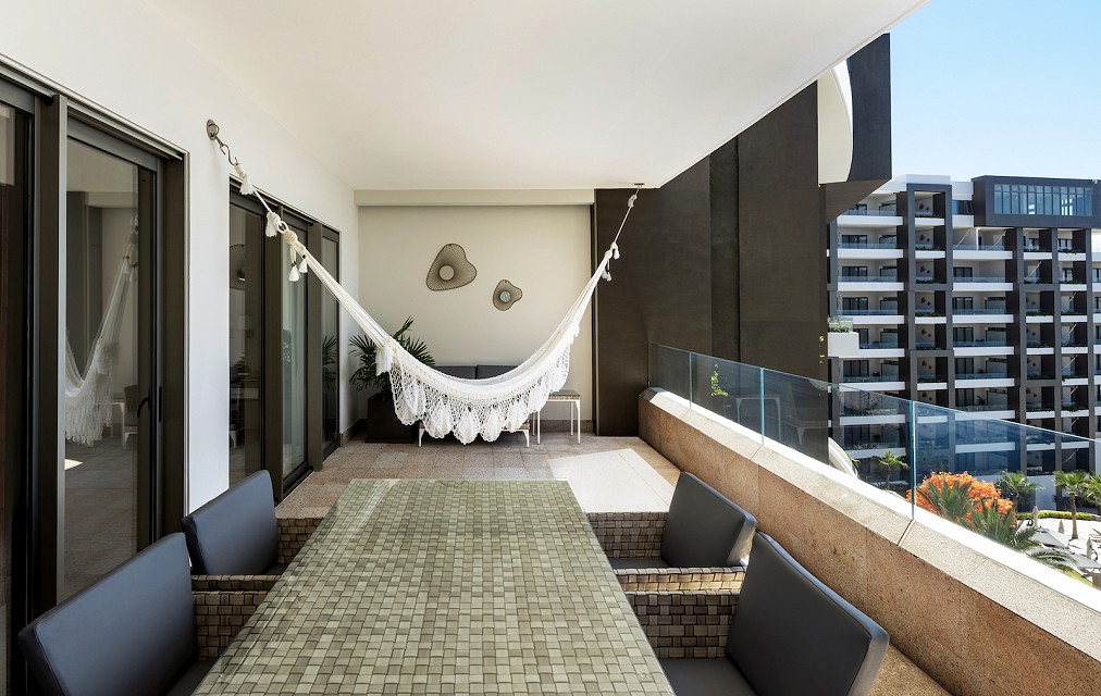 Modern balcony with a hammock, glass railing, and a dining table with gray chairs, overlooking urban buildings and greenery.