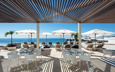 Rooftop terrace with white tables, wicker lounge chairs, and umbrellas overlooking the ocean. Palm trees and a slatted pergola provide shade.