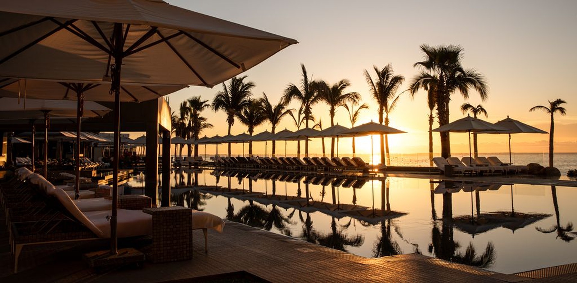 Sunset view of a tranquil poolside scene with palm trees, lounge chairs, and umbrellas reflecting on the water's surface.