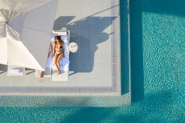 A woman in a bikini sunbathes on a poolside lounger under a large umbrella, surrounded by clear turquoise water.