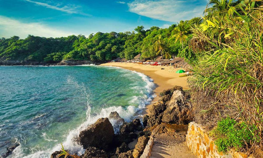 Idyllic tropical beach with golden sand, lush greenery, and sun loungers under straw umbrellas in puerto escondido