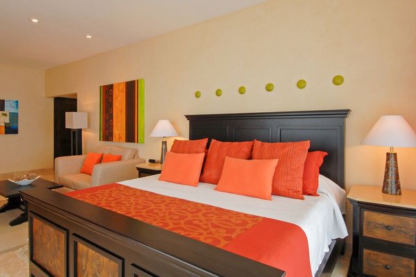 A cozy bedroom with a large bed featuring orange and red pillows and a matching runner, flanked by two lamps on wooden nightstands.