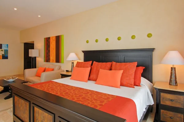A cozy bedroom with a large bed featuring orange and red pillows and a matching runner, flanked by two lamps on wooden nightstands.